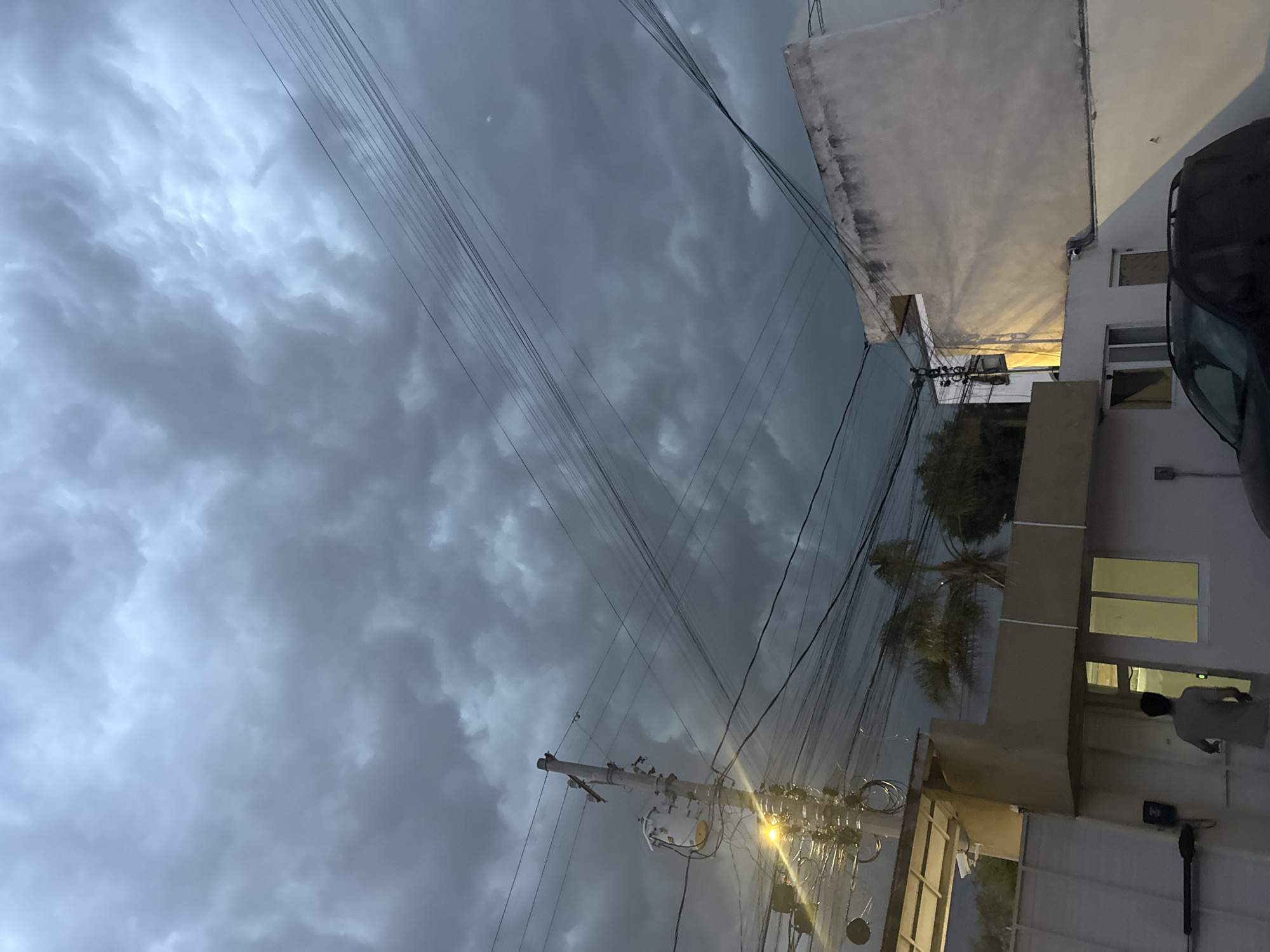 The image, taken in San Andrés Cholula, Mexico, depicts a cloudy, overcast sky filling most of the frame. Power lines crisscross the view of the sky. A building with windows emitting light stands on the right side of the image. A dark vehicle is partially visible in the top right corner, next to the building. Palm trees and other foliage are situated near the building. A utility pole with transformers and wires is visible in the lower center of the image, illuminated by a nearby light source. A person is visible through one of the building's windows.