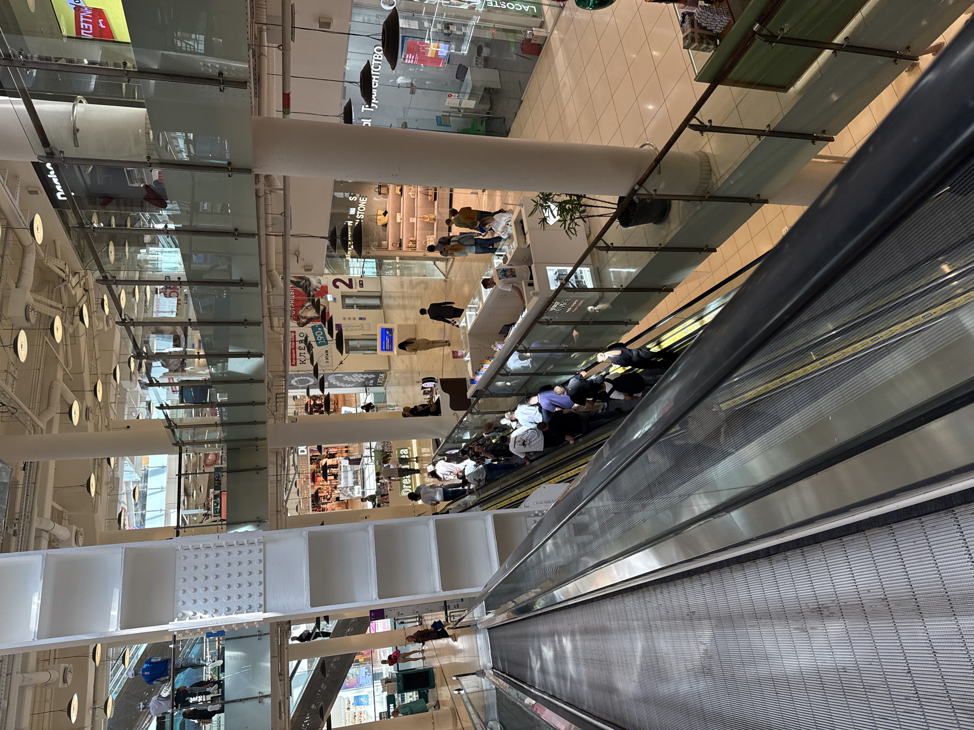 This image depicts the interior of a multi-level shopping mall in Khimki, Russia. The primary focus is on people using an escalator and a travelator. Several individuals are visible, some standing on the escalator, while others are walking or standing nearby on the mall's various levels. The architecture of the mall is modern, featuring glass railings, white pillars, and a bright, tiled floor. Shops are visible on different floors, with some signage indicating brands like Lacoste.
The activity is the everyday movement of shoppers within the mall. The lighting suggests it is daytime. There are no specific weather cues visible as it is an indoor scene. The overall emotion conveyed is neutral, reflecting a typical day in a commercial space. Visible text includes brand names and directional indicators within the mall, such as "LACOSTE," "Drea a," "STONE," "КЛЁВО," and floor numbers like "1 ЭТАЖ" and "2 ЭТАЖ." The presence of Cyrillic script confirms the Russian location.