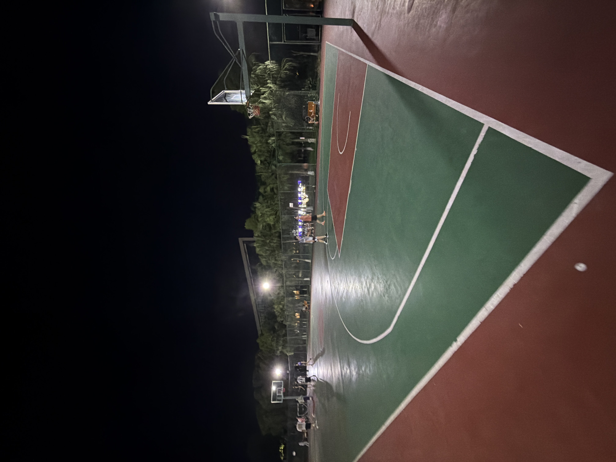 The image shows an outdoor basketball court at night. The court has a red and green surface with white lines. There are several people on the court, some playing basketball and others observing. The court is lit by overhead lights, and trees and buildings are visible in the background. The scene appears to be in a recreational area or park. It is not possible to determine the specific location within Jiangjin, China from the image alone, but it is consistent with an urban park or sports facility within the city.