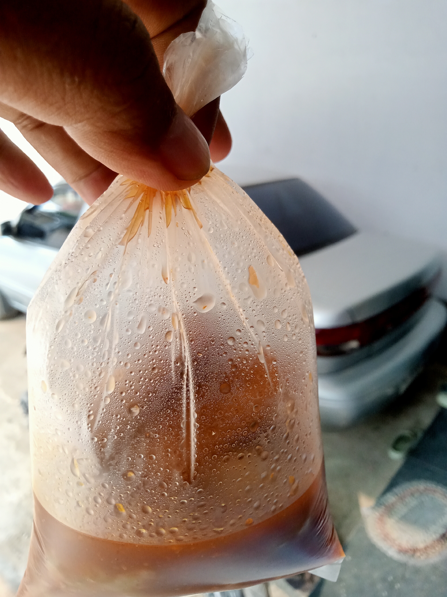 A close-up shot captures a hand holding a clear plastic bag filled with a dark brown liquid, likely a beverage. The bag is condensation-covered, with numerous small water droplets clinging to its surface. The liquid inside appears to have some solid particles suspended in it, possibly ice or ingredients. In the blurred background, a silver car is visible, suggesting an outdoor setting, potentially a street or parking area. The overall scene implies a casual, everyday moment, possibly involving the purchase or consumption of a drink. The specific location within Simo, Indonesia, cannot be determined from the visual information alone.