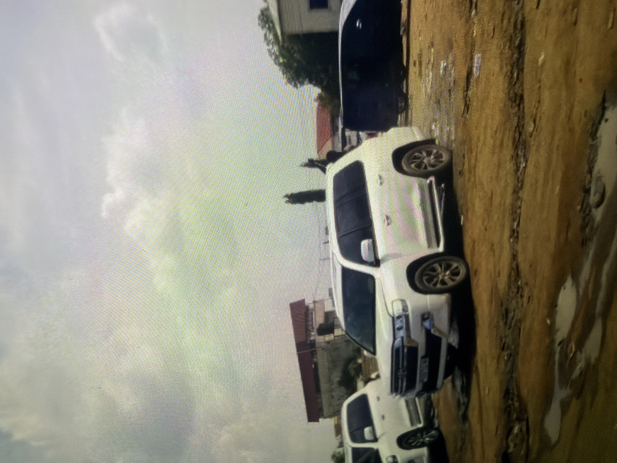 In Bukuru, Nigeria, a cluster of vehicles is parked on a dirt lot, under a cloudy sky. The vehicles include two white SUVs, one of which is prominently featured in the foreground. The front of this white SUV is turned towards the camera, revealing its grille and headlights. Behind it, another white SUV is visible, parked to its left. A dark-colored vehicle, possibly an SUV or van, is parked to the right of the foreground white SUV. In the background, a building with a reddish-brown roof and concrete walls stands on a slight incline. Another building, appearing lighter in color, is situated further back to the right, partially obscured by trees. The ground surface is rough and uneven, with patches of dry earth and puddles of water, indicating a recent rainfall or an area prone to accumulation. The overall scene depicts an outdoor parking area with a backdrop of urban structures and natural elements.