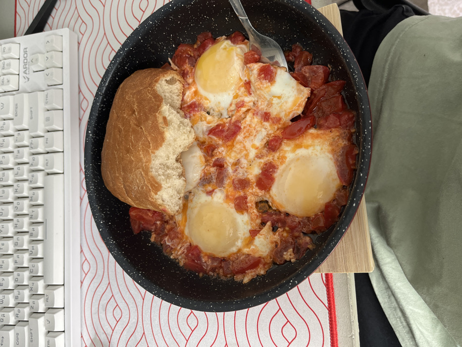 An indoor scene in Zainsk, Russia, displays a black cooking pan, speckled with white, containing four fried eggs nestled in a vibrant red tomato-based sauce with visible pieces of red pepper or sausage. A substantial, light brown piece of bread, with a distinct bite mark on its side, rests on top of the food. A silver spoon is partially submerged on the right side of the pan. The pan sits on a light-colored wooden trivet, which is placed on a white desk mat featuring a repeating red wave pattern. In the background, a white mechanical keyboard with Russian Cyrillic and Latin characters is visible, bearing the "ARDOR" brand logo in its upper right corner. Below the pan, a light green fabric, consistent with clothing, is present. No individuals are explicitly visible, but the arrangement suggests a person is consuming the meal at a desk or table.