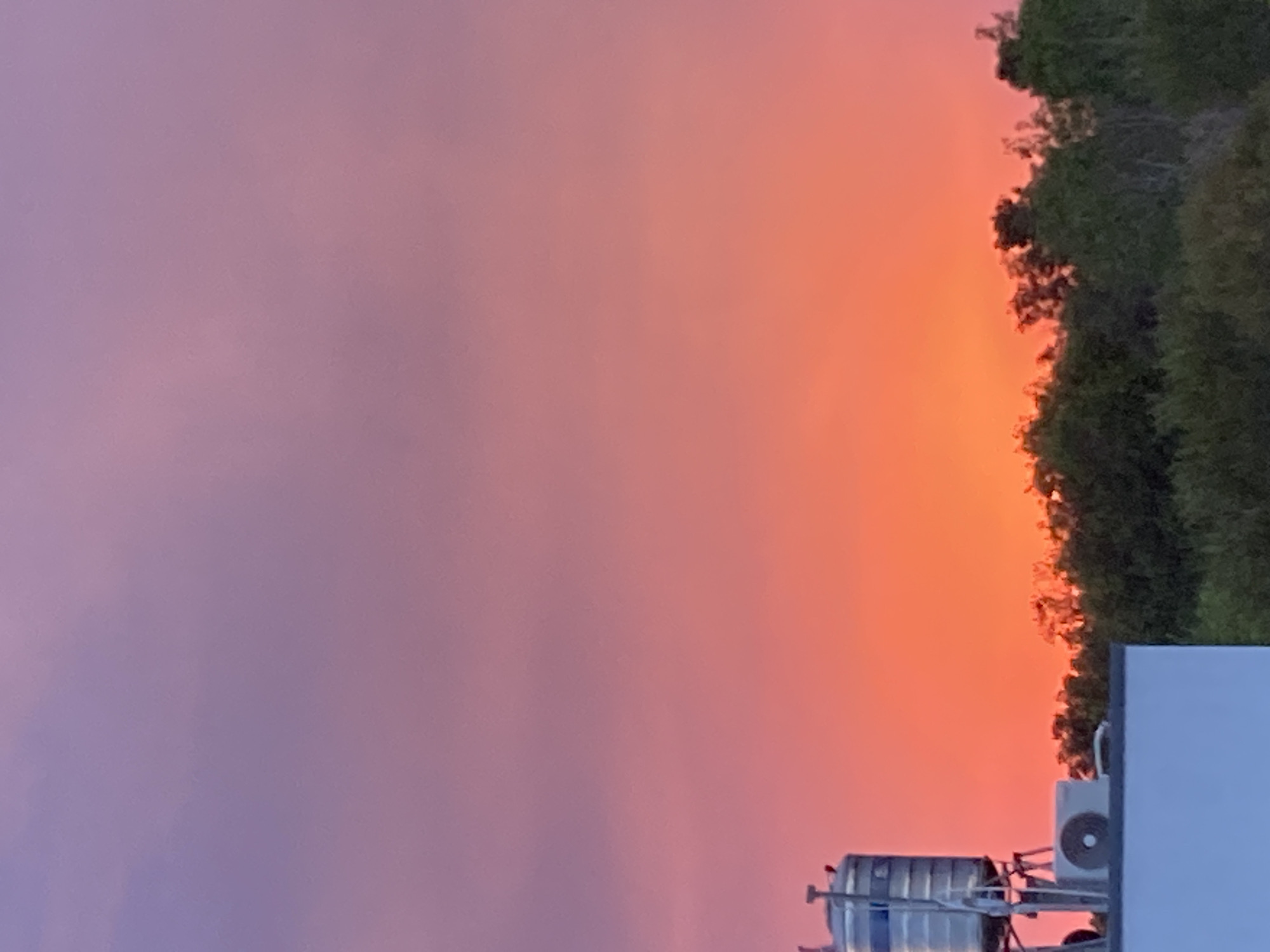The image shows a vibrant sunset sky in Mũi Né, Vietnam. The sky transitions from purple on the left to orange on the right. Silhouetted trees are visible against the orange sky on the right side of the image. In the foreground, there is a structure with metal tanks, an air conditioning unit, and a white wall. The image is rotated counterclockwise.