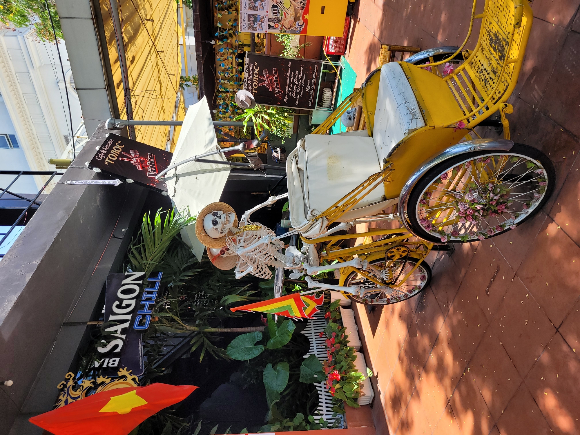 The image shows a yellow vehicle resembling a rickshaw with white cushions and a skeleton wearing a straw hat in the driver's seat. The rickshaw's wheels are decorated with flowers. Behind the rickshaw is a restaurant with a black sign that reads "SAIGON CHILL" and "BIA." There is also a red and yellow Vietnamese flag. The area appears to be outdoors on a tiled surface, possibly a sidewalk or patio. Additional signage visible includes "Cafe & Karaoke 'ГОЛОС'" and "Voice." The location is in Nha Trang, as indicated on one of the signs.