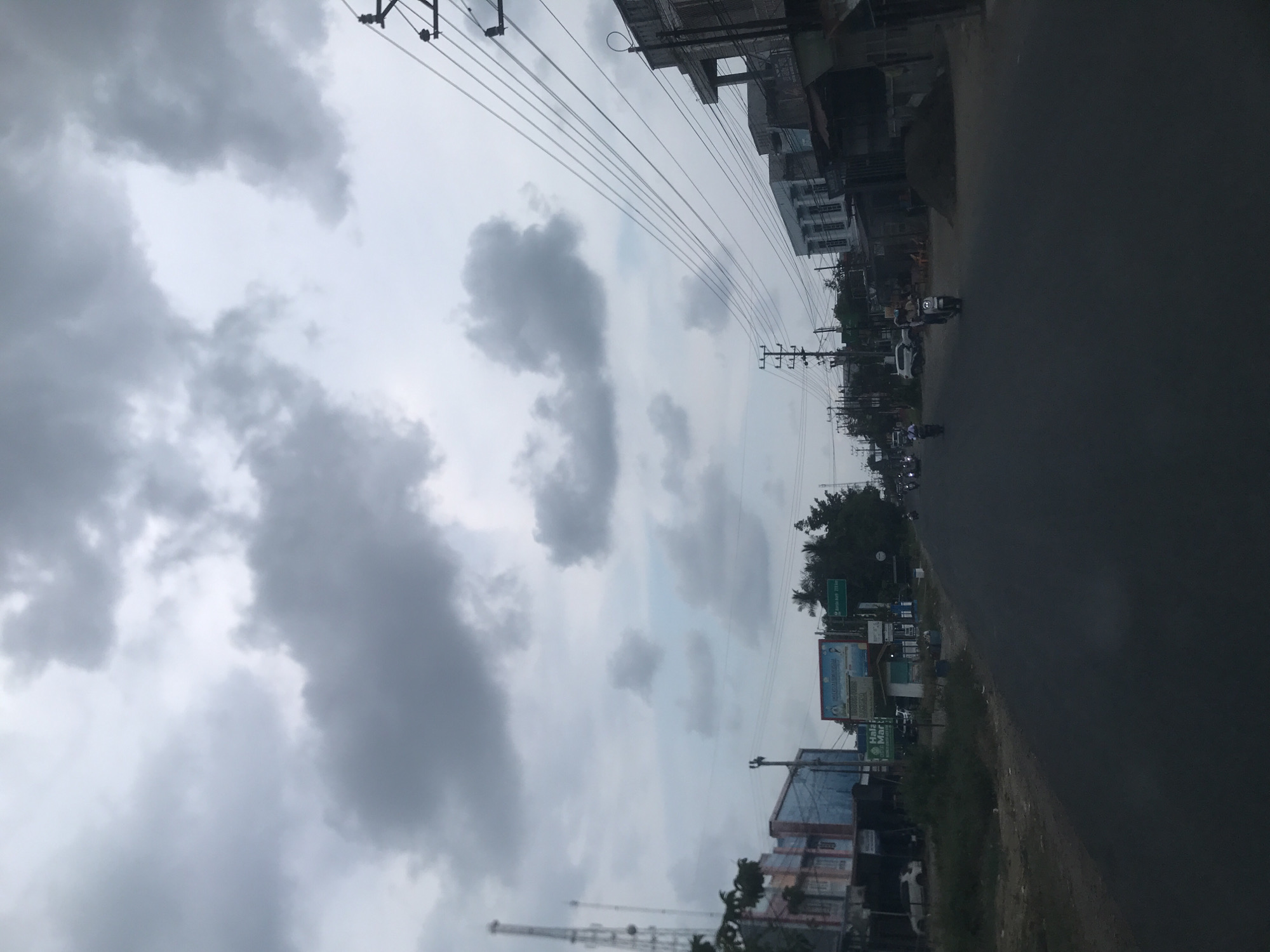 The image shows a street scene in Lhokseumawe, Indonesia, under a cloudy sky. The road occupies the right side of the frame and leads into the distance where vehicles, including motorbikes, are visible. Buildings line the right side of the road. Power lines stretch across the sky. Nearer the viewer, a sign can be seen. A transmission tower rises in the lower center of the image.