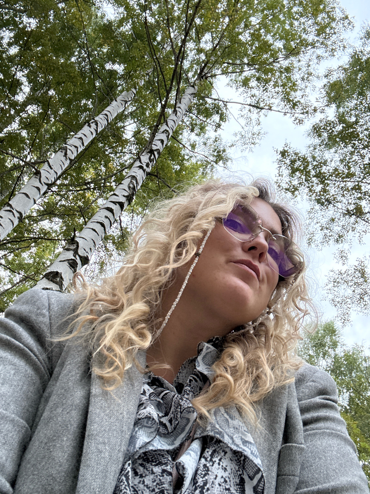 A low-angle, close-up, outdoor shot features a Caucasian woman with curly blonde hair and light skin. She is wearing a gray blazer over a black and white patterned scarf. She has on rimless sunglasses with a purple tint and a beaded string attached to them, which loops around her ear. The woman is looking upwards. In the background, two birch trees with distinctive white bark and black markings are prominent, with green foliage visible above and around them. The sky appears overcast. The setting is likely a park or wooded area in Chaikovsky, Russia, given the presence of birch trees common in the region.