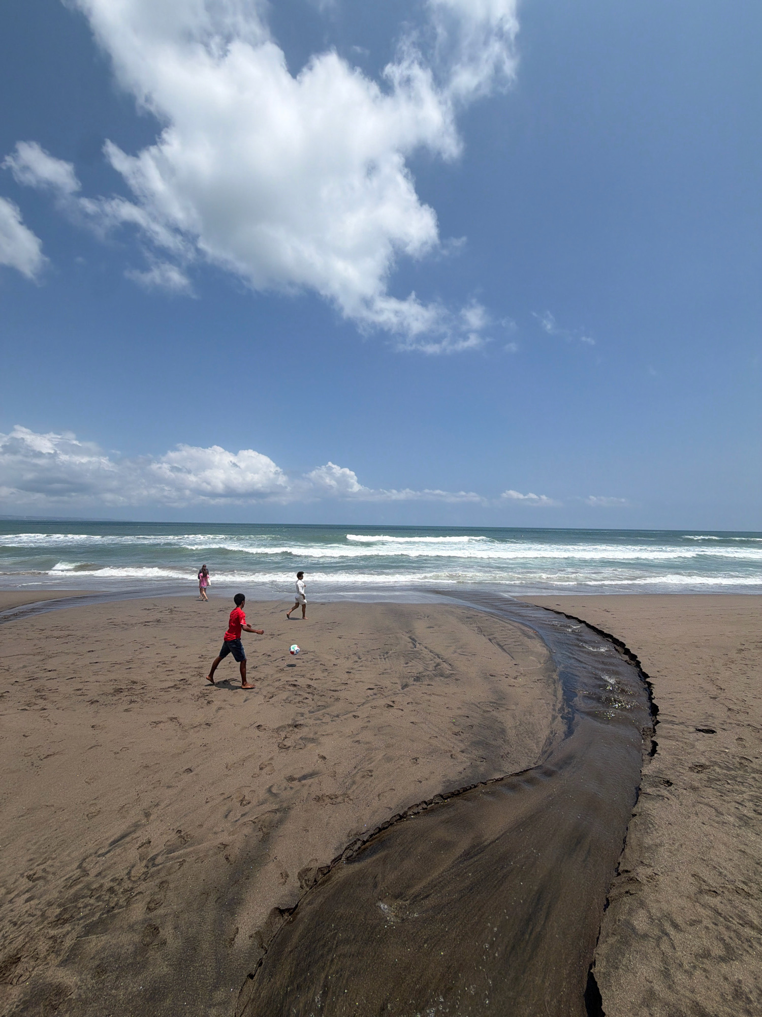 In North Kuta, Indonesia, a beach scene unfolds under a bright, blue sky dotted with fluffy white clouds. The sand is a warm brown, marked by the footprints of visitors and a dark, winding stream that flows towards the ocean. The ocean itself is a deep blue-green, with white-capped waves rolling onto the shore. In the middle ground, three individuals are engaged in a game of soccer. One person, wearing a red shirt and blue shorts, is mid-kick, sending a white soccer ball into the air. Another person, in white shorts, stands nearby, observing the game. Further back, a third person in a pink shirt walks along the water's edge. The overall impression is one of a relaxed and active day at the beach.