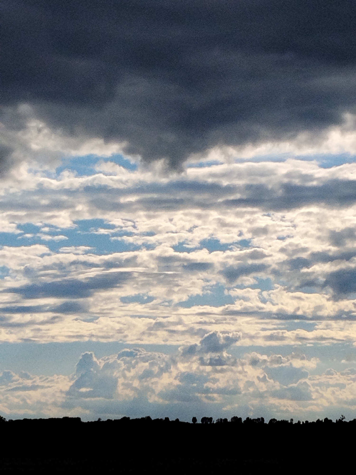 This is a photograph taken in Ustyvytsya, Ukraine, depicting a dramatic sky filled with a variety of clouds. The upper portion of the image is dominated by a large, dark, ominous cloud formation, suggesting impending weather. Below this, a series of lighter, layered clouds stretch horizontally across the frame, interspersed with patches of blue sky. The lowest layer of clouds is composed of more voluminous, fluffy formations. The bottom of the image shows a silhouetted landscape of trees and possibly distant structures against the bright sky, indicating a horizon line. The overall impression is one of a vast, active sky over a rural or semi-rural area.