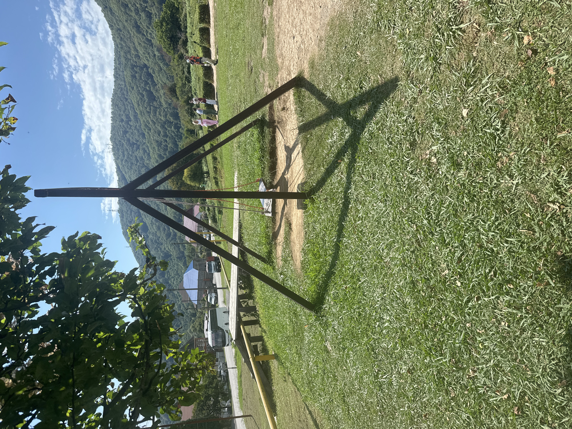 The image depicts an outdoor scene in the city of Bolshoy Kichmai, Russia. In the foreground, a wooden swing set with a simple seat hangs from a pyramidal frame. The frame casts a distinct shadow on the grassy ground. To the left of the swing set, a yellow metal railing is visible, partially obscured by green foliage from a tree overhead. In the middle ground, a group of people are gathered, appearing to be a family or friends, observing something. Behind them, a path leads to a landscaped area with hedges. Further in the background, a paved road runs past several buildings, including a white bus and other vehicles. The scene is set against a backdrop of lush green hills and mountains, under a bright blue sky with scattered clouds. The environment suggests a park or recreational area, possibly in a rural or suburban part of the city.