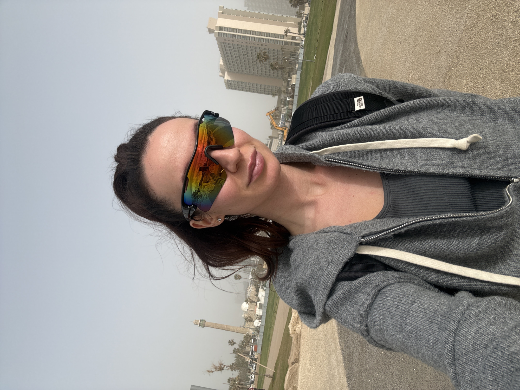 A woman in a gray zip-up hoodie and colorful sunglasses is taking a selfie outdoors. She is standing on a paved area, with buildings and greenery visible in the background. The location is Tel Aviv, Israel. A tall building and other structures are reflected in the woman’s sunglasses. The scene appears hazy.