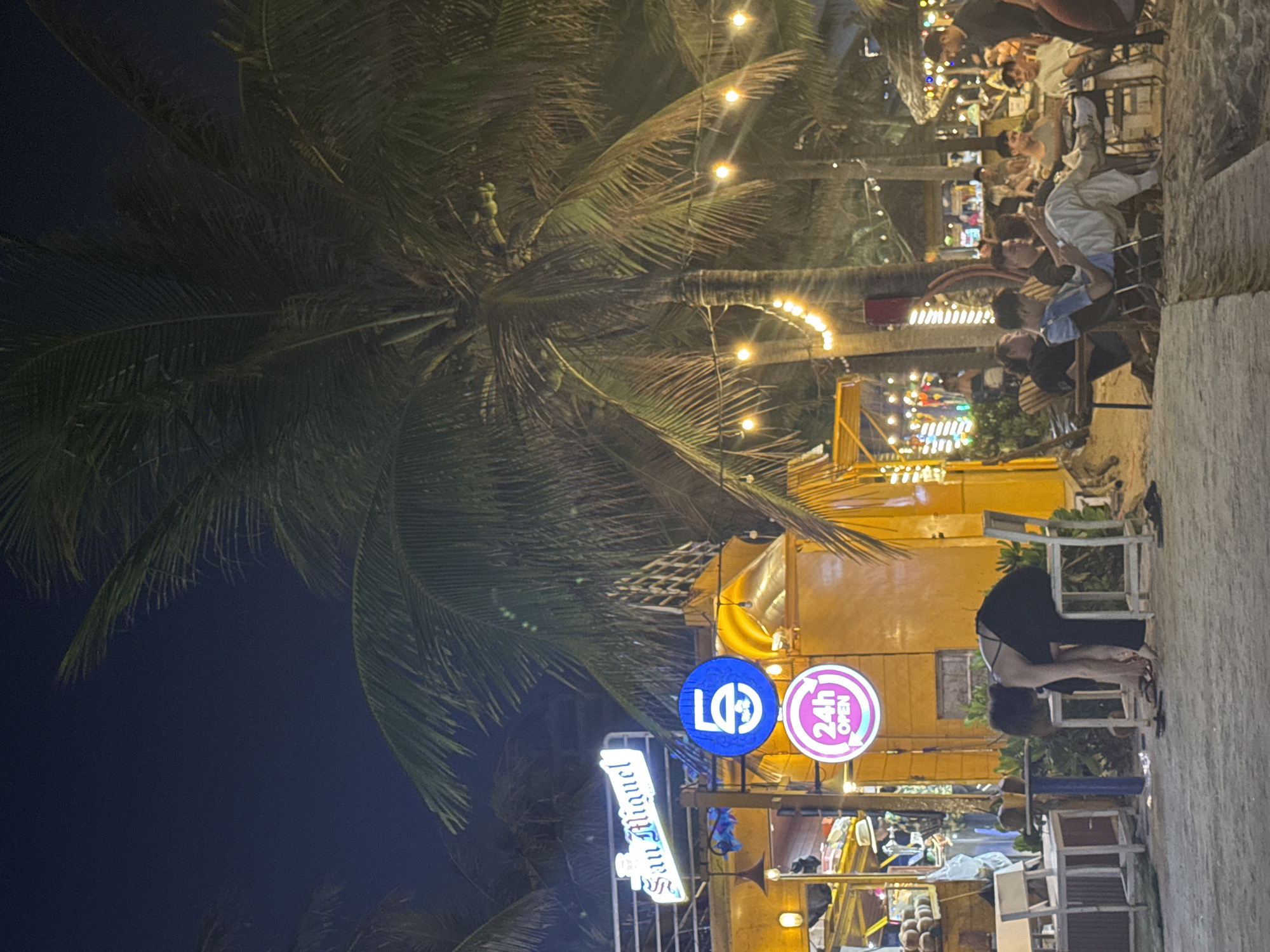 At night, a bustling outdoor scene is captured. A tall palm tree dominates the left side of the frame. String lights twinkle above, illuminating people seated at tables. The foreground contains illuminated signage for "San Miguel" and an "Open 24 hours" sign. A person is bending over near some chairs. The setting appears to be a beachside venue with patrons enjoying the evening. It is located in the city of Đà Nẵng, Vietnam.
