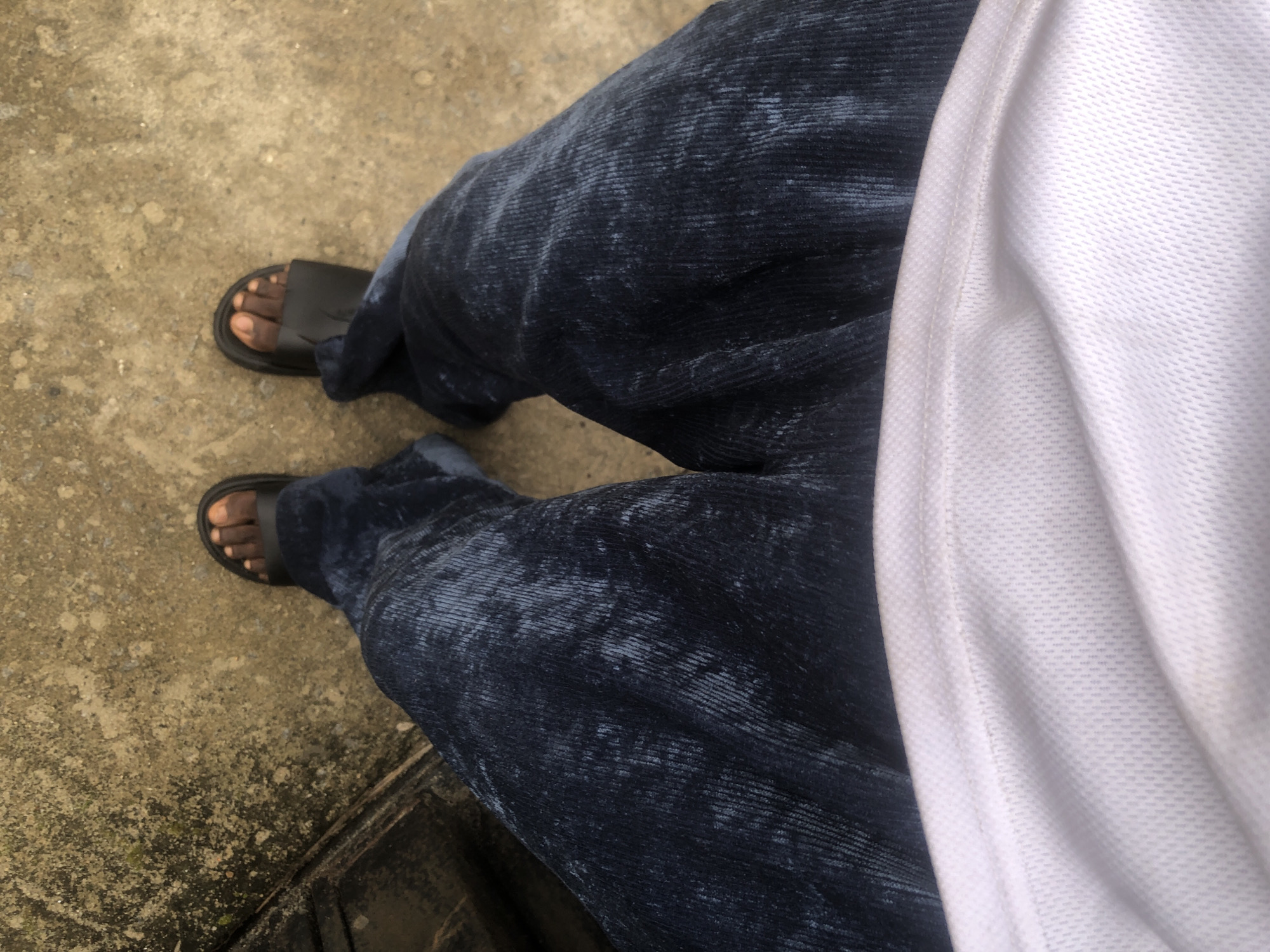 The image shows a person wearing dark blue corduroy pants and black slide sandals. The person is standing on a concrete surface. The scene is likely outdoors, possibly in a street or walkway in Ado, Nigeria. The lighting suggests it is daytime. The perspective is from above, looking down at the person's legs and feet. The concrete surface appears somewhat weathered and textured. A portion of a white garment, possibly a shirt, is visible at the bottom of the frame.