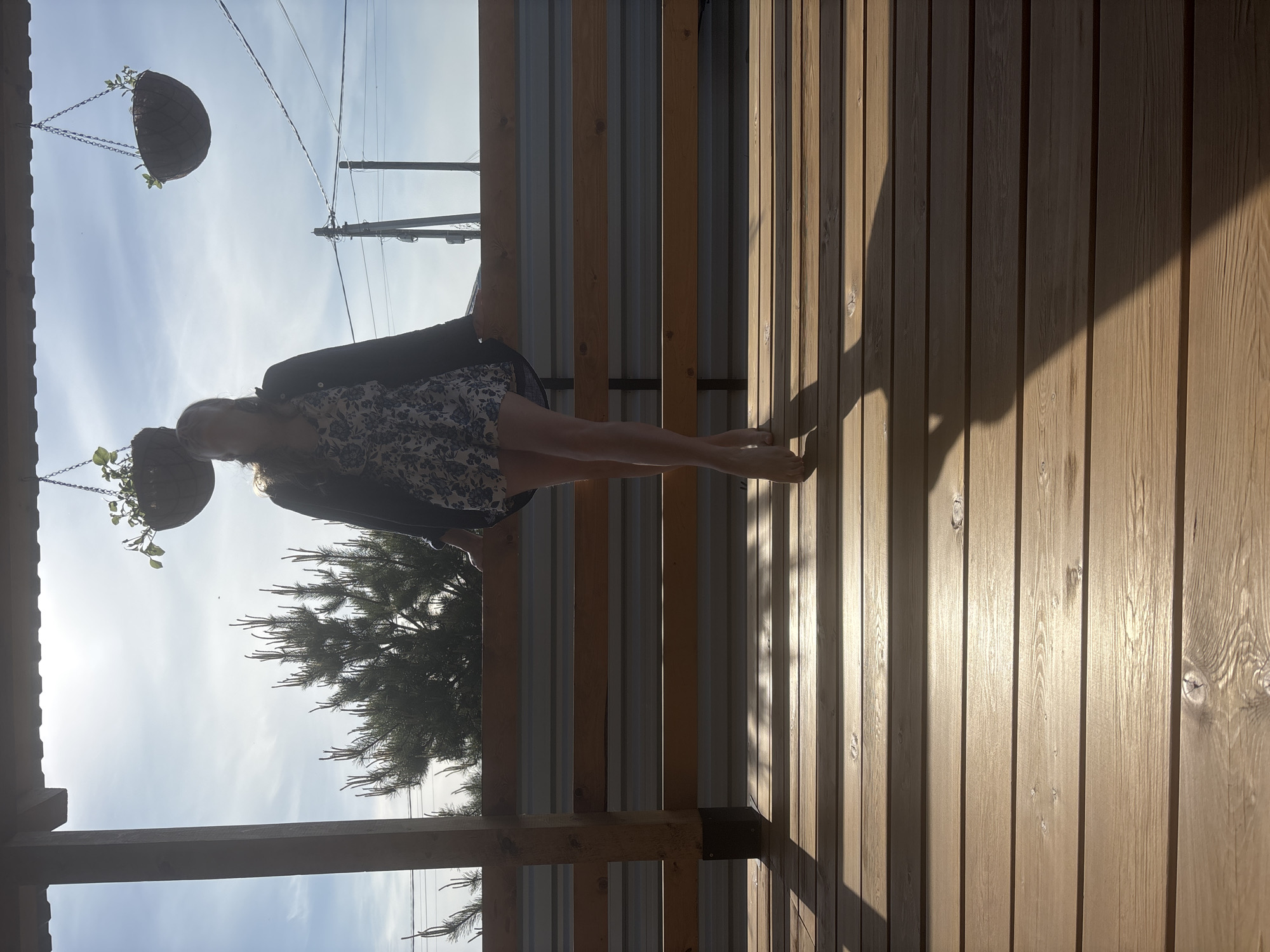 Here's a description of the image:
The image shows a young woman standing on a wooden deck, seemingly in a backyard or outdoor area. She is facing away from the camera and toward the outside of the deck. She is wearing a short, floral-patterned dress and a dark-colored jacket. Her feet are bare.
The deck is made of parallel wooden planks. To the left of the woman is a pine tree, visible against a bright sky. Above her, hanging from the roof or structure above the deck, are two hanging baskets with plants. Power lines are visible in the background.
The light suggests it's daytime, with shadows cast by the structure and the woman. The location may be Chaikovsky, Russia, although this cannot be confirmed from the image itself.