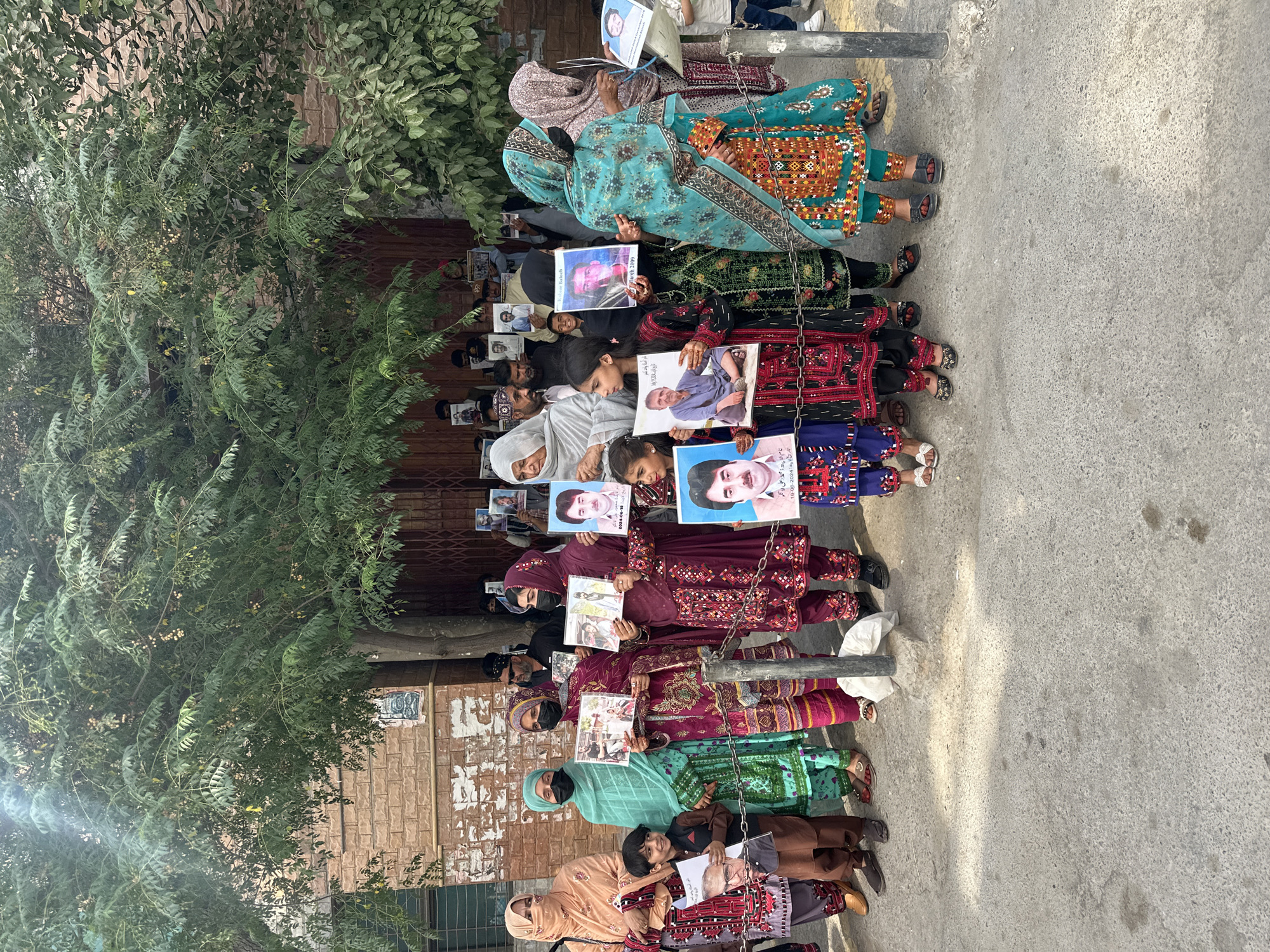 A group of people, mostly women, are standing in a line holding up photographs. They appear to be protesting or mourning. The event is taking place outdoors on a paved surface, possibly a sidewalk, in what looks like an urban setting. The group is located in the city of Quetta City Tehsil, Pakistan. Many of the people are wearing traditional Pakistani clothing. The photographs they are holding feature images of men. Trees and a building are visible in the background.