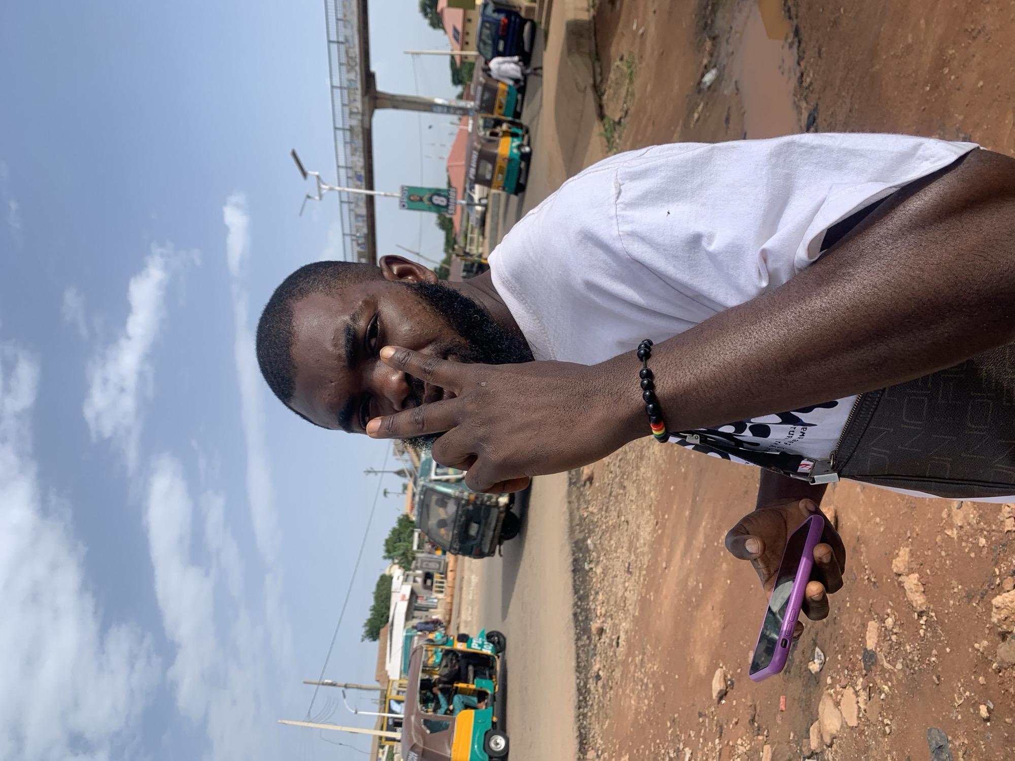 A man with dark skin, a beard, and short hair is standing on a dirt road. He is wearing a white t-shirt, a black crossbody bag with a patterned strap, and a bracelet on his left wrist with red, yellow, and green beads. He is holding a purple smartphone in his left hand and has his right hand raised, with his index and middle fingers extended in a peace sign. He is looking directly at the camera.
In the background, there is a busy street scene. Several auto-rickshaws, also known as "Keke Napep," are visible on the road, along with other vehicles. There are buildings and shops lining the street, and a pedestrian overpass can be seen in the distance. The sky is partly cloudy. The location appears to be a street in Gura Top, Nigeria, given the presence of Keke Napep vehicles and the overall urban environment. The scene is illuminated by natural daylight.