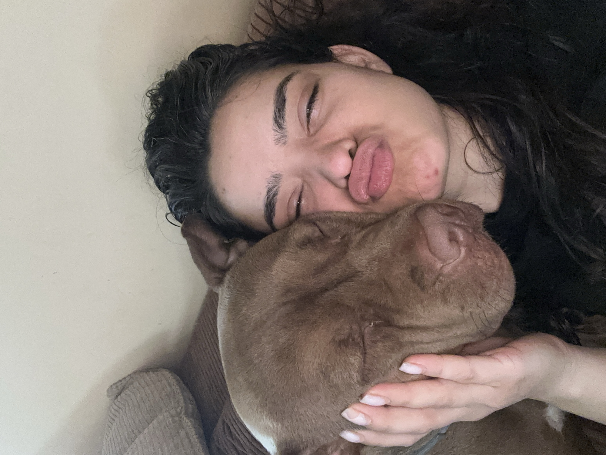 The image shows a close-up, overhead shot of a woman and a dog. The woman has dark, curly hair and is lying down, her eyes closed and her lips puckered. She is resting her head on the dog's head. The dog is a pit bull with brown fur and is also lying down, its head nestled against the woman. A hand, presumably the woman's, is gently resting on the dog's cheek. The background is a plain, light-colored wall. The overall mood appears to be affectionate and relaxed, suggesting a moment of intimacy between the woman and her pet. The lighting suggests it's daytime. There is no visible text in the image.