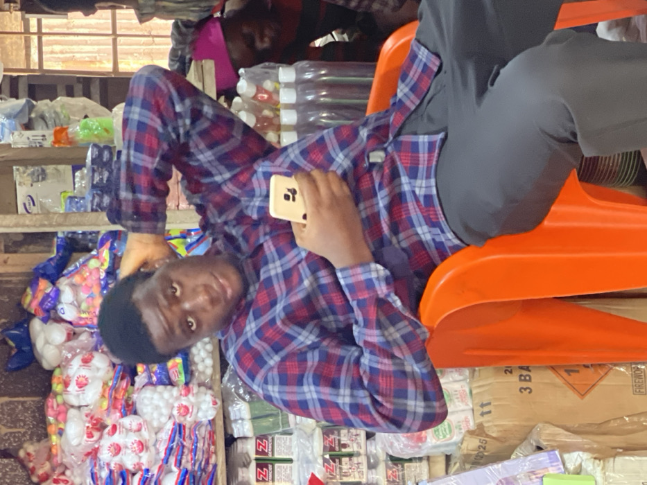 A man in a plaid shirt and grey pants leans back in an orange plastic chair, holding a phone. He appears to be in a shop or market, surrounded by various items for sale including packages of marshmallows, bottled drinks, and boxes labelled "FIREWORKS." Another person is visible in the background, also in a plaid shirt. The setting suggests a busy, informal commercial environment.