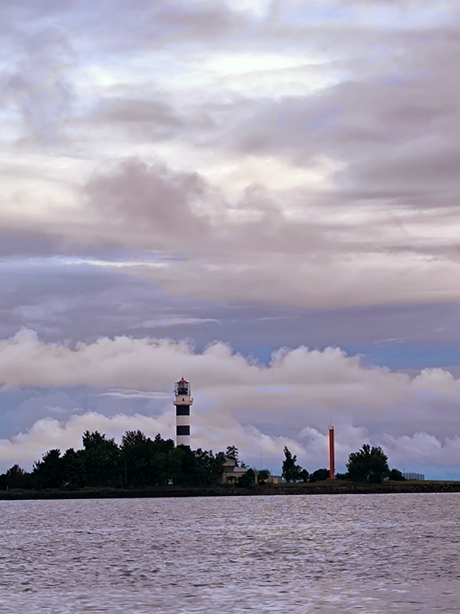 This is a photograph taken from the water, showing a view of the coast of Riga, Latvia. The foreground is dominated by the choppy surface of a body of water, appearing as a muted lavender or pink color due to the lighting conditions. In the middle ground, a strip of land is visible, covered with dense green trees. Prominently featured on this land are two lighthouses. The larger, more central lighthouse is tall and slender, with alternating black and white horizontal stripes. It has a red lantern room at its apex. To the right of this lighthouse, a smaller, bright red cylindrical lighthouse stands. The background is filled with a dramatic sky of swirling clouds. The clouds are a mixture of light grey, white, and soft purples and blues, suggesting either sunrise or sunset. The overall atmosphere is slightly overcast but serene.