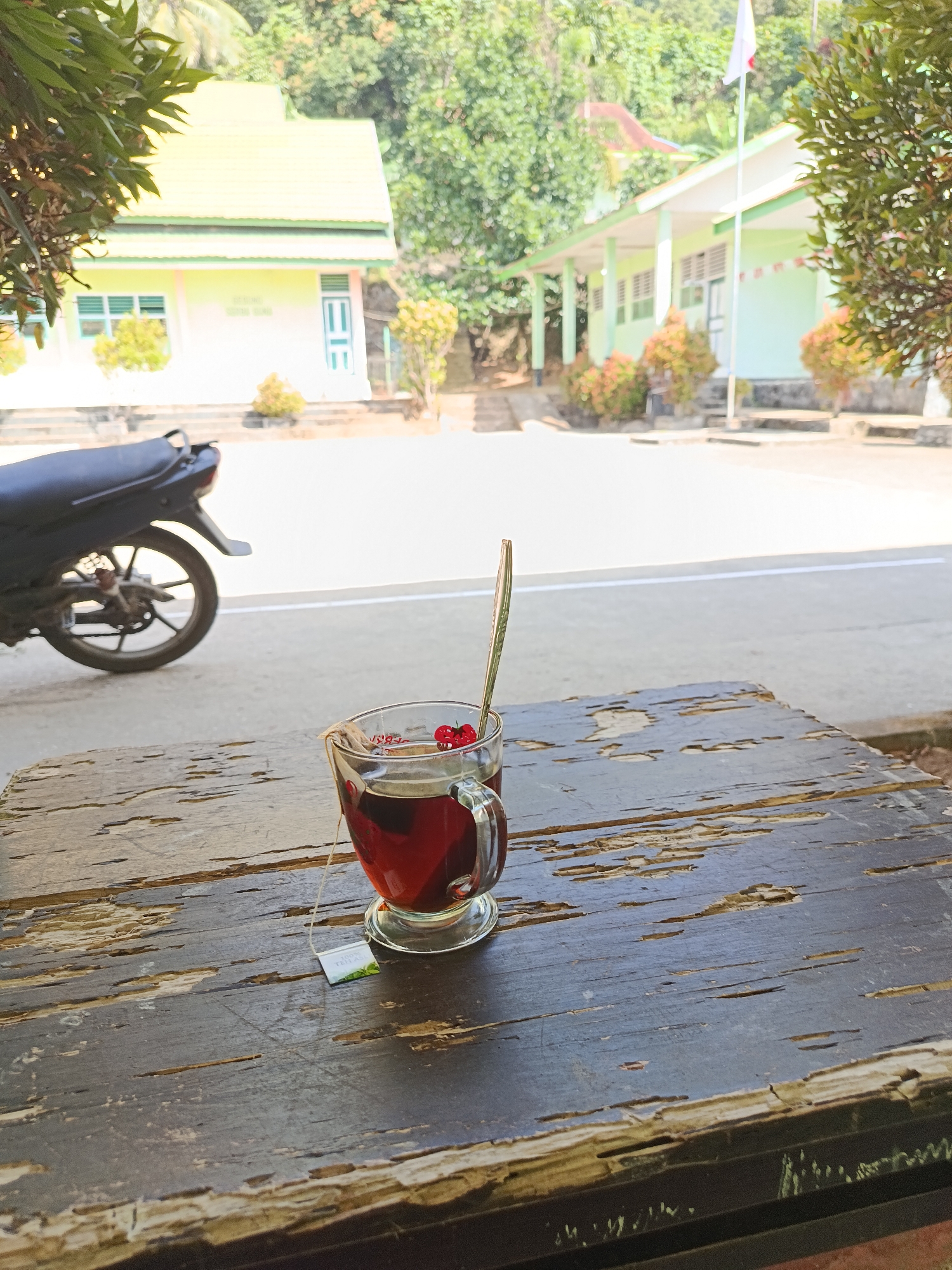 The image captures a close-up of a glass cup of tea, complete with a tea bag and a spoon adorned with two red berries, resting on a weathered wooden table. The background reveals a bright, outdoor scene with buildings that appear to be part of a school or administrative complex in Mansalean, Indonesia. The buildings have light-colored walls and distinctive green roofs. A motorcycle is parked in the middle ground, to the left of the tea cup. Lush green foliage and trees surround the structures, indicating a tropical or subtropical environment. The overall atmosphere is serene and casual, possibly depicting a moment of rest or refreshment at an outdoor location.