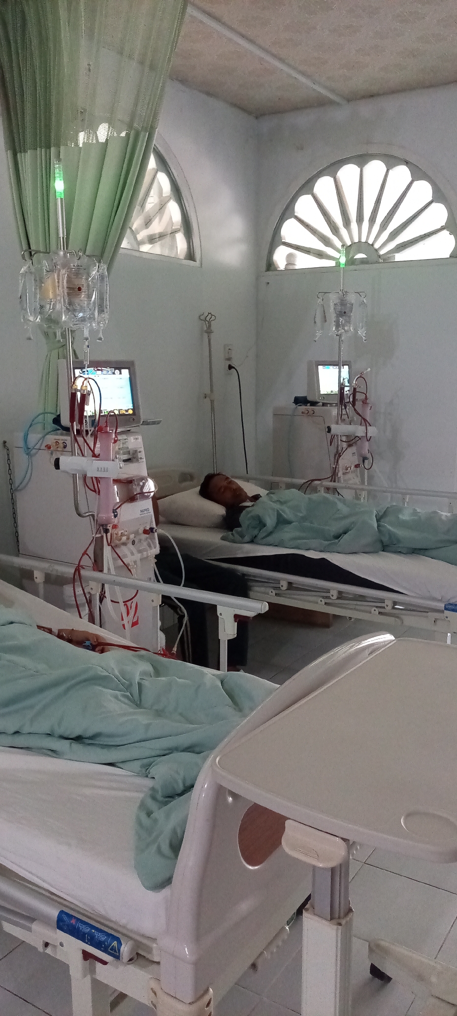 The image depicts a hospital room with two beds set up for dialysis. In the foreground, a patient is covered by a light blue sheet, with only their arm, connected to medical tubing, visible. A dialysis machine stands next to the bed, with bags of fluid hanging from an IV pole and a monitor displaying data. In the background, another patient lies in a bed, also covered by a blue sheet, with a similar dialysis setup. The room has arched windows with a fan-like design. The overall impression is one of a clinical environment focused on medical treatment. The image is tagged with Lhokseumawe, Indonesia, suggesting this is a view of a dialysis unit in that city.