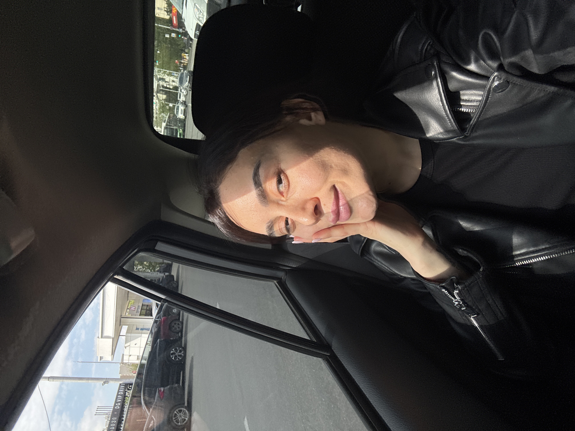 An upward-tilted, slightly rotated, medium close-up shot captures a woman inside a car, bathed in bright sunlight. The woman, positioned on the right side of the frame, is wearing a black leather jacket over a dark shirt. She rests her chin on her right hand, showcasing manicured nails with light polish. Sunlight casts a distinct diagonal shadow across her face, highlighting her left eye and the left side of her face, while the right side remains in shadow. She has dark hair, defined eyebrows, and a gentle smile, looking directly towards the camera with a relaxed expression.
Through the car windows on the left and upper left, an outdoor urban scene is visible, appearing rotated. The background shows parked cars, a utility pole, and parts of buildings, including a white structure. Greenery and what appears to be a distant traffic light are also discernible. A dark sign with white text, partially obscured, reads "DG VIP DRIVE". The interior of the car, including door panels and window frames, is visible in dark tones, contrasting with the bright exterior and the illuminated parts of the woman. The strong sunlight and clear skies indicate it is daytime. The scene is set in Tbilisi, Georgia.