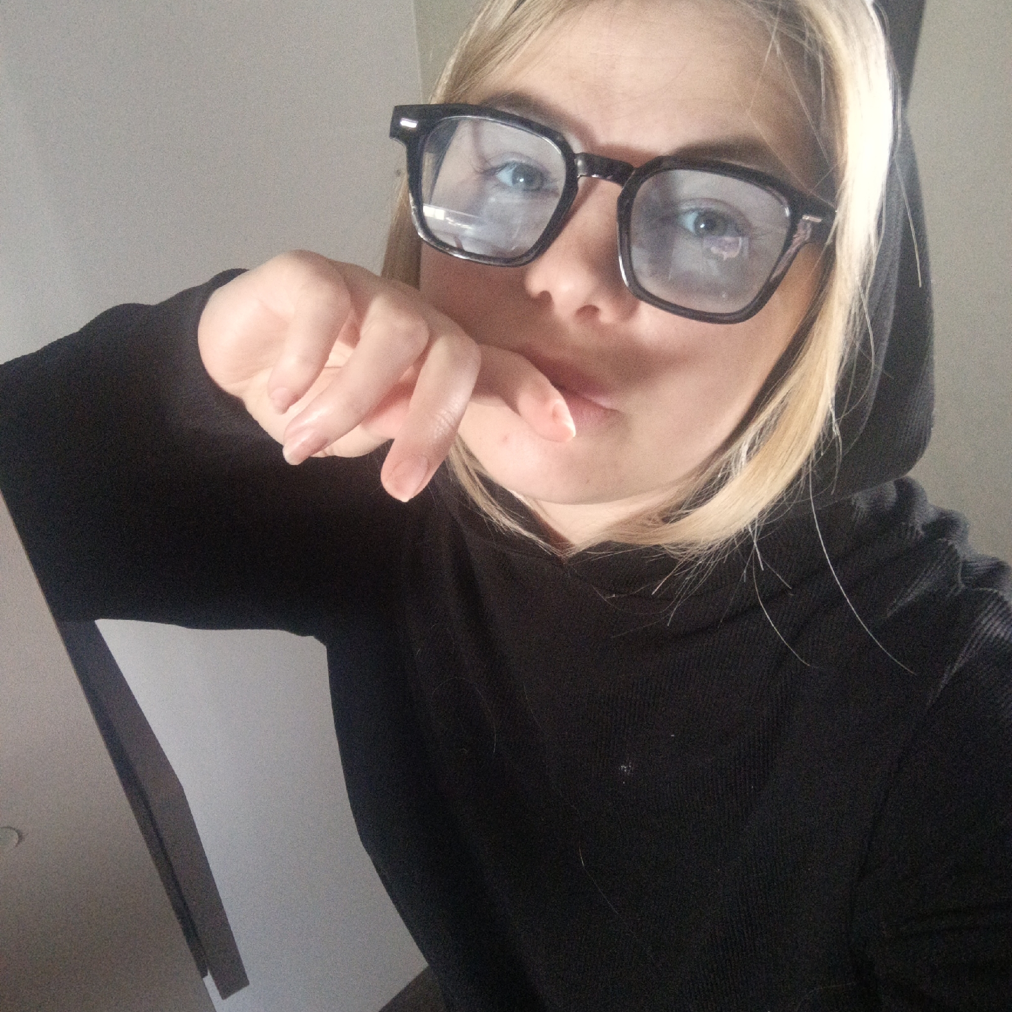 A candid, medium-close-up shot features a person with blonde hair, fair skin, and blue eyes, wearing black, thick-rimmed glasses and a black hooded sweatshirt. The subject is looking directly towards the camera. Their right hand is raised, with fingers gently touching their chin and lower lip, creating a contemplative or casual pose. Reflections are visible on the lenses of the glasses, indicating light sources. Loose strands of blonde hair fall around the face and over the shoulders of the black sweatshirt. The background is a plain, light-colored wall, with a dark, possibly wooden, structure or piece of furniture visible on the lower left side of the frame. The image has a slightly grainy texture. There is no visible text. The setting appears to be indoors.