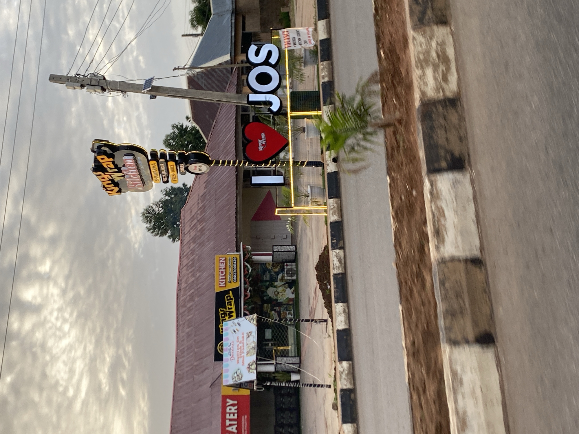 The image captures a street scene in Bar Jirgi Summa, Nigeria, featuring a prominent sign for "Kings Wrap Kitchen" and an "I ❤️ JOS" display. The "Kings Wrap Kitchen" sign is illuminated and lists menu items like Shawarma, Pizza, Burger, Fresh Juice, and Grills, along with "Free Wi-Fi." Adjacent to it, another sign advertises "Little Desserts" with images of waffles and mini pancakes. The "I ❤️ JOS" display consists of large illuminated letters and a heart symbol with the "Kings Wrap" logo inside. The buildings behind the signs appear to be shops with awnings. A utility pole with electrical wires and some solar panels is visible on the right side of the frame. The street in the foreground has a paved road and a concrete curb with black and white markings. The sky is cloudy. There are no people visible in the image.
