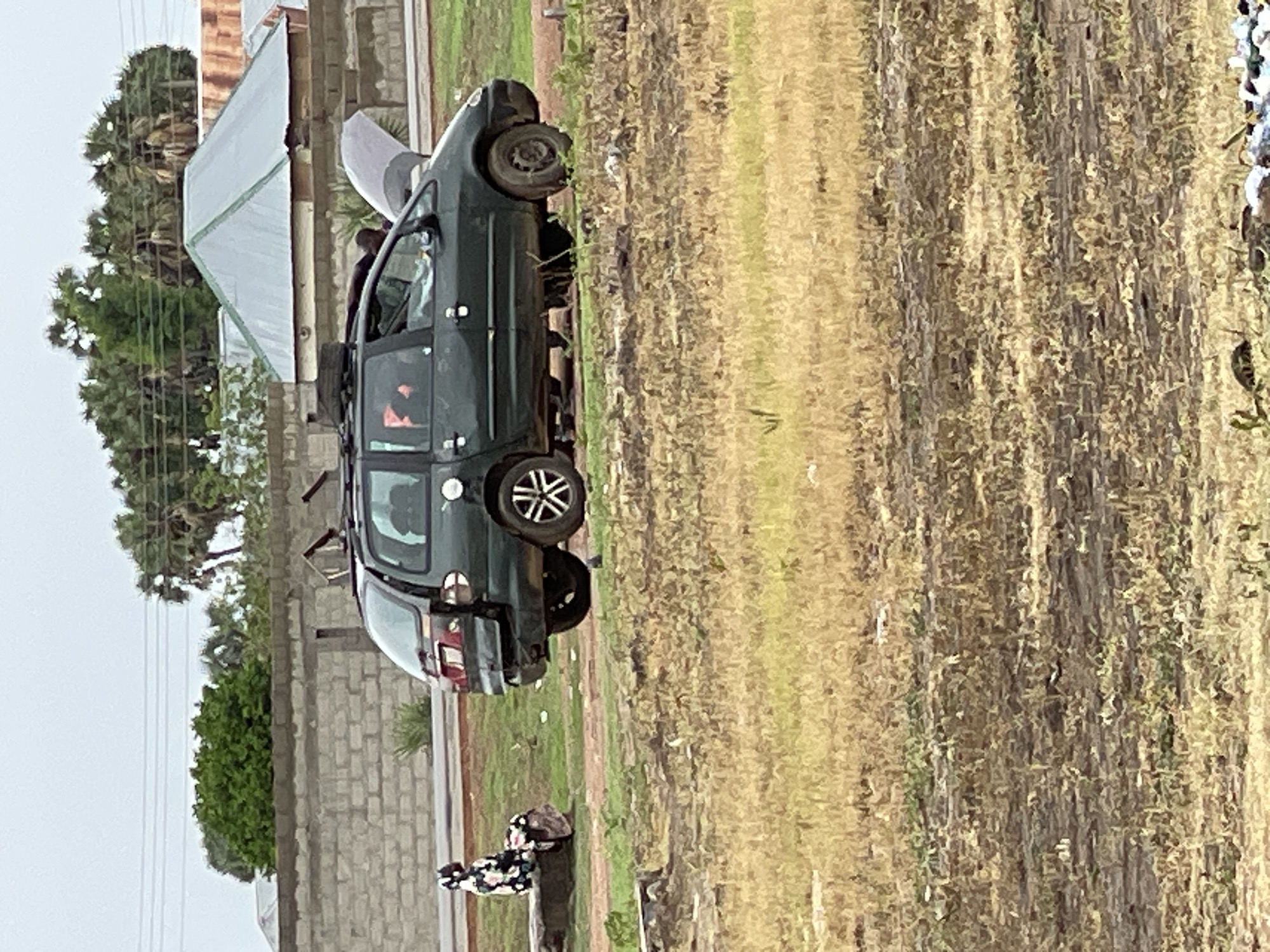 An outdoor, medium shot captures a dark green minivan with its hood up, seemingly undergoing maintenance, in a semi-urban or rural environment in Wai A, Nigeria. The minivan is parked on a dirt and grass area, with its rear slightly damaged. A person in a patterned headscarf and dress is seated on a concrete block to the left of the vehicle, facing away from the camera. In the background, a stone wall and buildings with corrugated metal roofs are visible, along with a cluster of palm trees. The ground in the foreground is dry and covered with sparse vegetation. The sky is overcast.