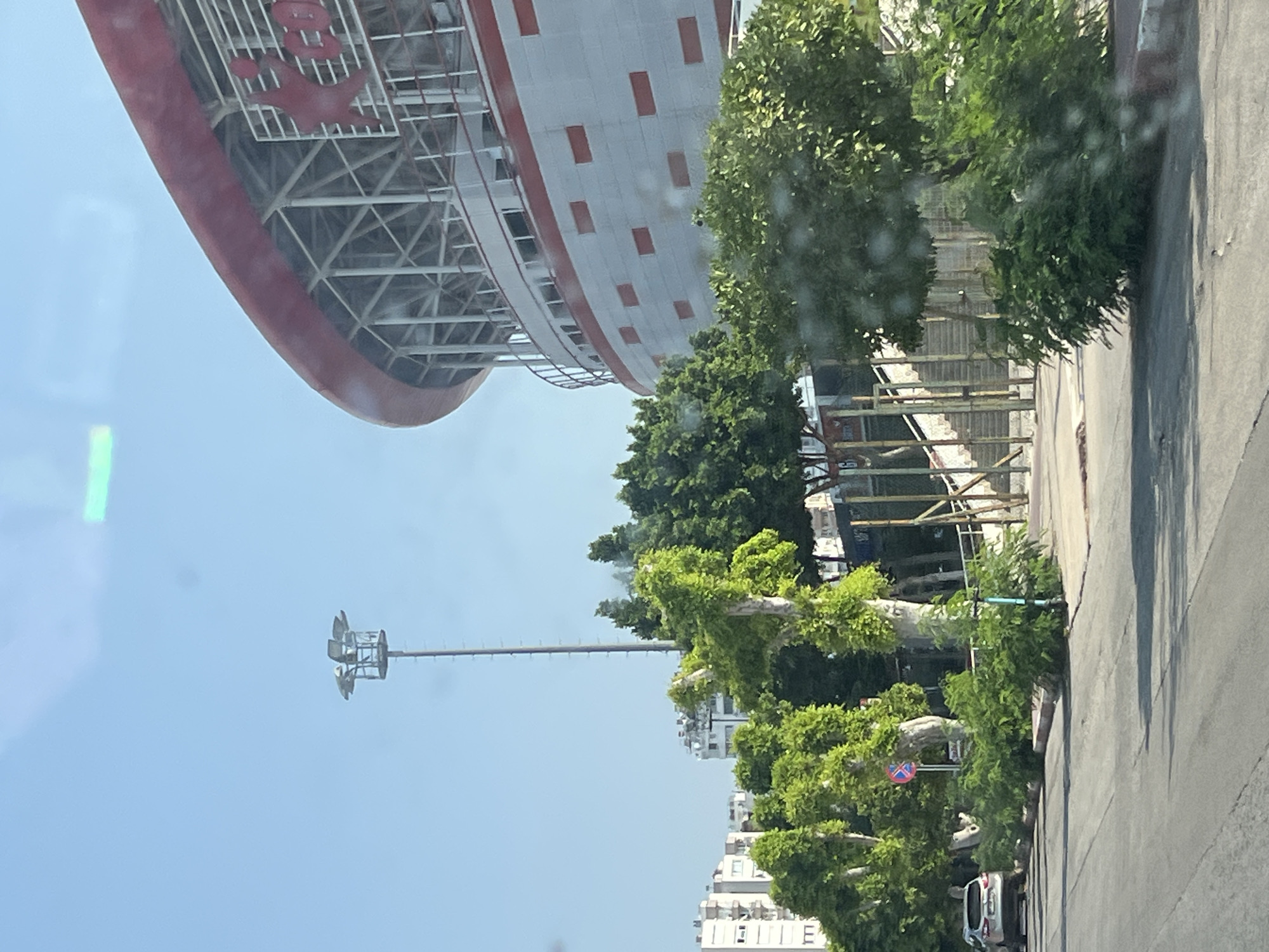 The image depicts a street scene in Antalya, Turkey, with a large, modern stadium dominating the right side of the frame. The stadium features a curved, red-and-white facade with a visible steel support structure. In the background, several residential buildings can be seen. The foreground shows a paved road with a light-colored car parked on the left side. Lush green trees and vegetation line the street, with some trees appearing to be covered in ivy. A tall lamppost with multiple lights stands prominently in the mid-ground. The sky is clear and blue, with some lens flare visible. The overall impression is one of a sunny day in an urban environment.