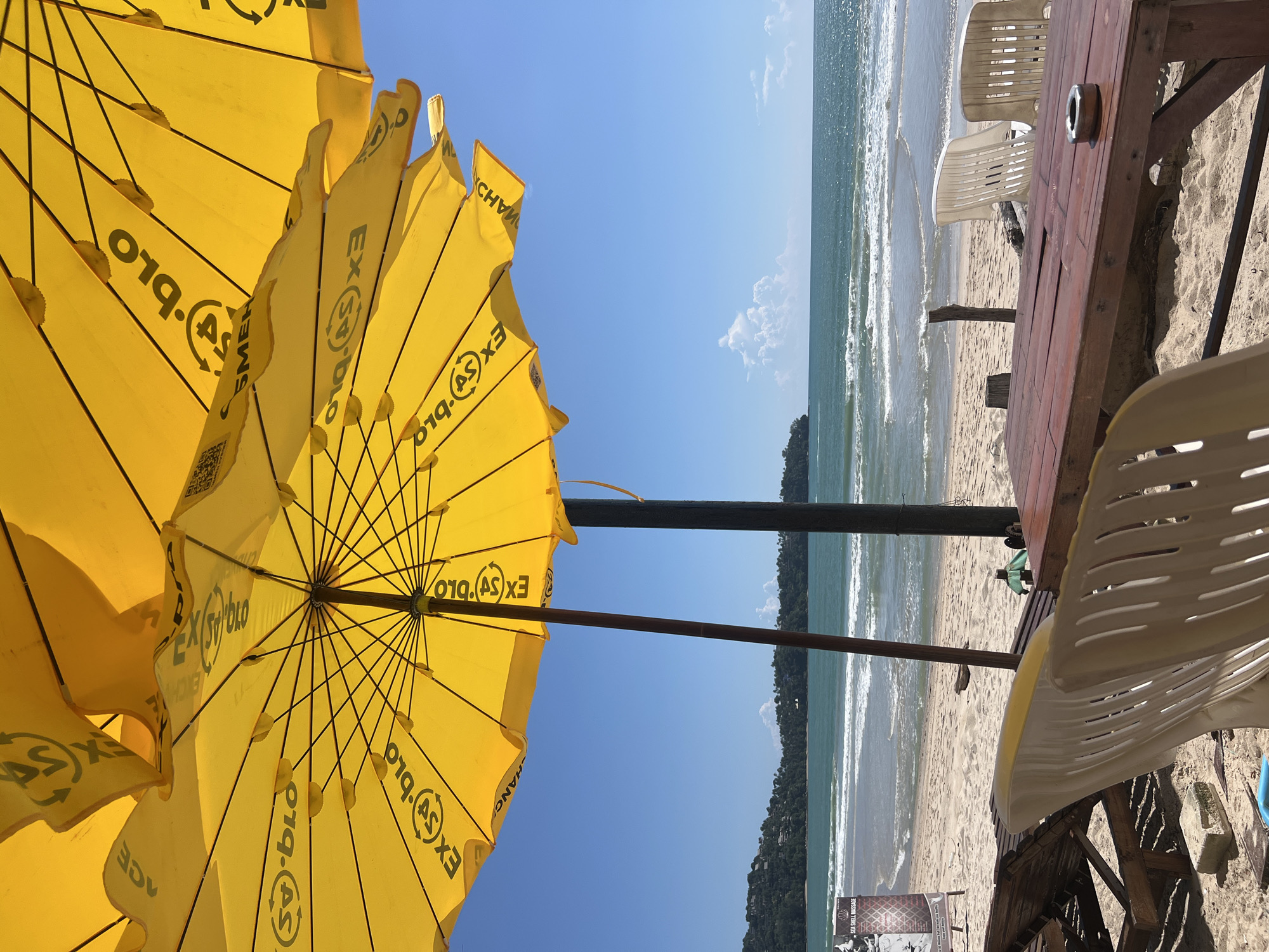 The image depicts a beach scene at Choeng Thale, Thailand, on a clear, sunny day. In the foreground, yellow beach umbrellas are prominently displayed, casting shadows and creating a vibrant overhead canopy. One umbrella's interior, with its black metal frame and fabric spokes, is clearly visible. The fabric of the umbrellas features repeating text, including "EX(SY)BLO" and "ACE," along with circular logos.
Beyond the umbrellas, a sandy beach stretches out, leading to the turquoise ocean. Gentle waves are breaking on the shore, and the water appears calm. In the middle ground, there is a wooden picnic table with several white plastic chairs arranged around it. An ashtray is visible on the table. Further back, several wooden lounge chairs are set up on the sand. A sign for "Sea Shell Massage" is visible on the left side of the image. The background features lush green hills covered in trees, forming the coastline. The sky is a bright, clear blue with a few scattered white clouds. No people are present in the frame, and the scene appears to be unoccupied at the time the photograph was taken. The overall impression is one of a serene and inviting beachside setting.