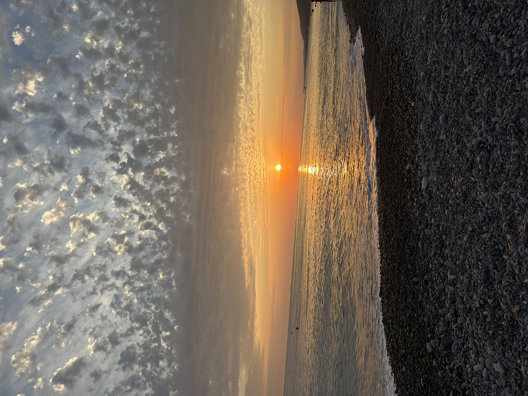 A wide shot shows a sunset over a pebble beach and the sea. The sun is setting on the left side of the frame, casting an orange glow across the sky and reflecting on the water. The sky is filled with clouds that are illuminated by the setting sun, creating a textured pattern. The sea is calm with small waves lapping at the shore. The beach is covered in dark, smooth pebbles. In the distance, a person is standing on the beach, silhouetted against the bright sky. The scene appears to be in a natural environment, likely a coastal area.