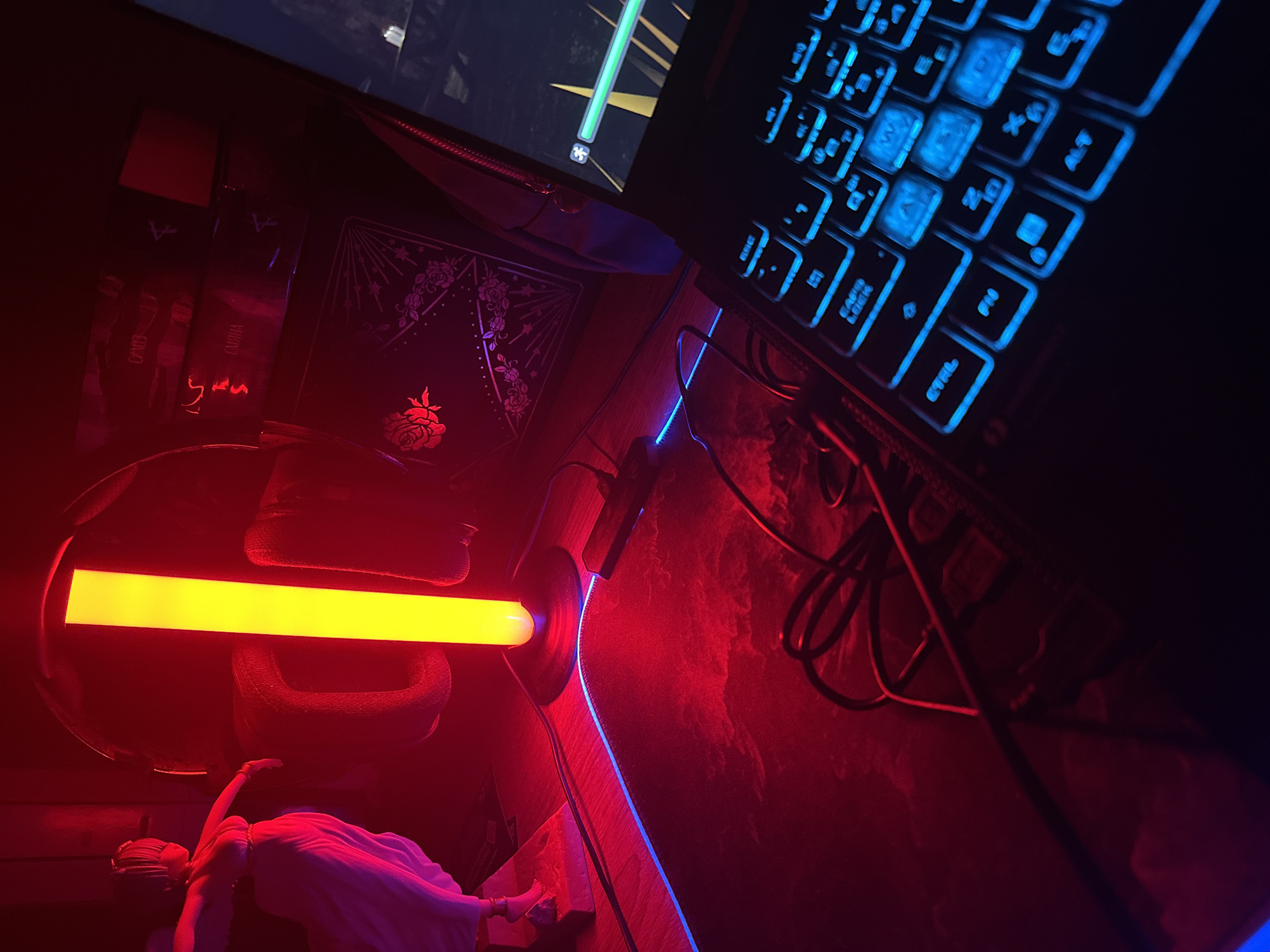 The image depicts a gaming setup illuminated by red and blue LED lighting. In the foreground, a black gaming mousepad with a cloudy pattern and blue LED trim is visible. A black laptop with illuminated blue keys is positioned on the right side of the mousepad, with its screen displaying a game interface with a green progress bar. To the left of the laptop, a black cable snakes across the mousepad. Behind the mousepad, a statue of a woman in a white toga stands on a wooden surface. A tall, vertical yellow LED light is situated next to the statue, casting a strong glow. In the background, stacked boxes with decorative patterns and what appear to be headphones are visible. The overall environment suggests a personal gaming space with a strong emphasis on atmospheric lighting.