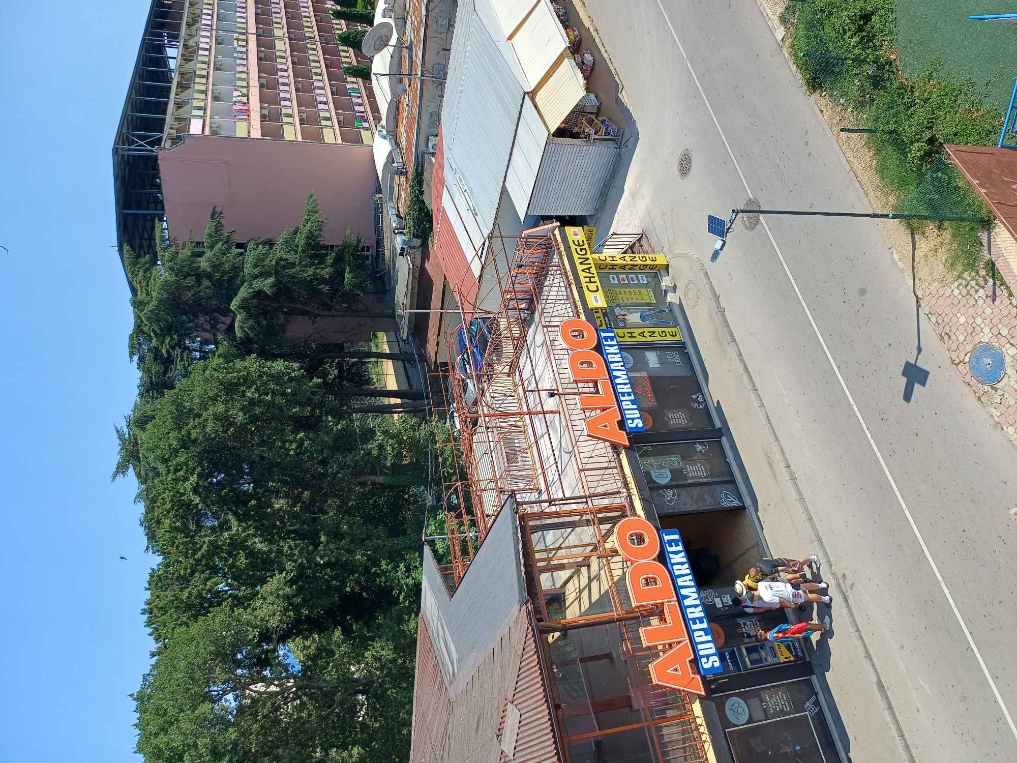 The image shows a sunny day in Varna, Bulgaria. In the foreground, a group of four young people are standing outside a store labeled "ALDO SUPERMARKET" and "CHANGE". The store has large orange lettering on its facade. To the right of the supermarket, there is a separate building with awnings, likely housing other shops. Behind these buildings, a large, multi-story pink building with many balconies is visible. Trees and greenery surround the area. The street in front of the supermarket has a solar-powered lamppost. The overall scene depicts a commercial area in a coastal city.