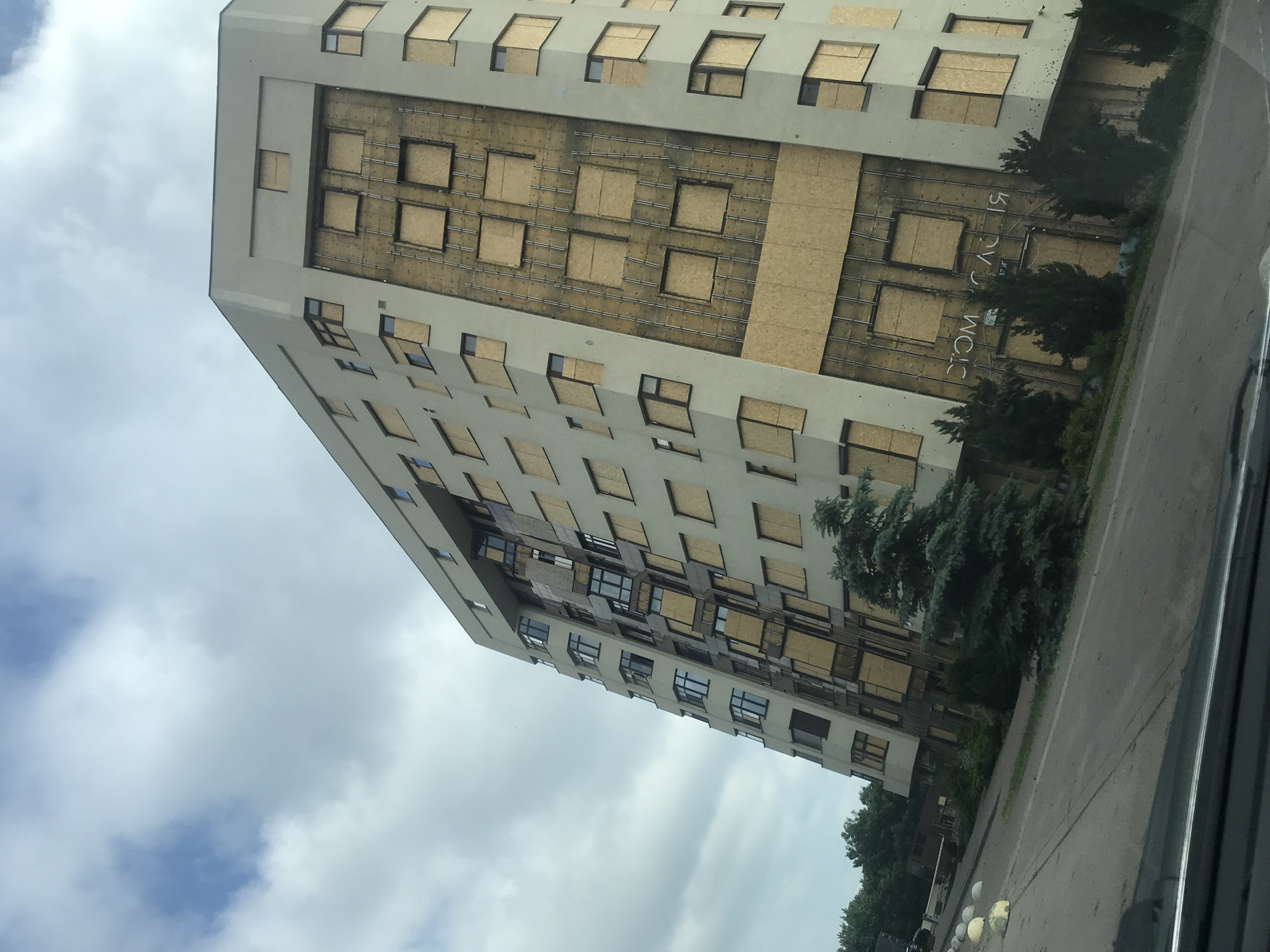 A tilted, long shot captures the exterior of a large, multi-story building that appears to be abandoned or under renovation, as many of its windows are boarded up with plywood. The building has a light-colored facade and a modern, geometric design. The boarded-up windows are arranged in a regular pattern, with some areas showing exposed structural elements. A sign with Cyrillic lettering is partially visible on the lower section of the building. The building is surrounded by a sidewalk, a road, and some trees. The sky above is overcast with scattered clouds. The overall impression is one of disuse and perhaps damage, given the boarded windows. The context provided places the scene in Kharkiv, Ukraine.