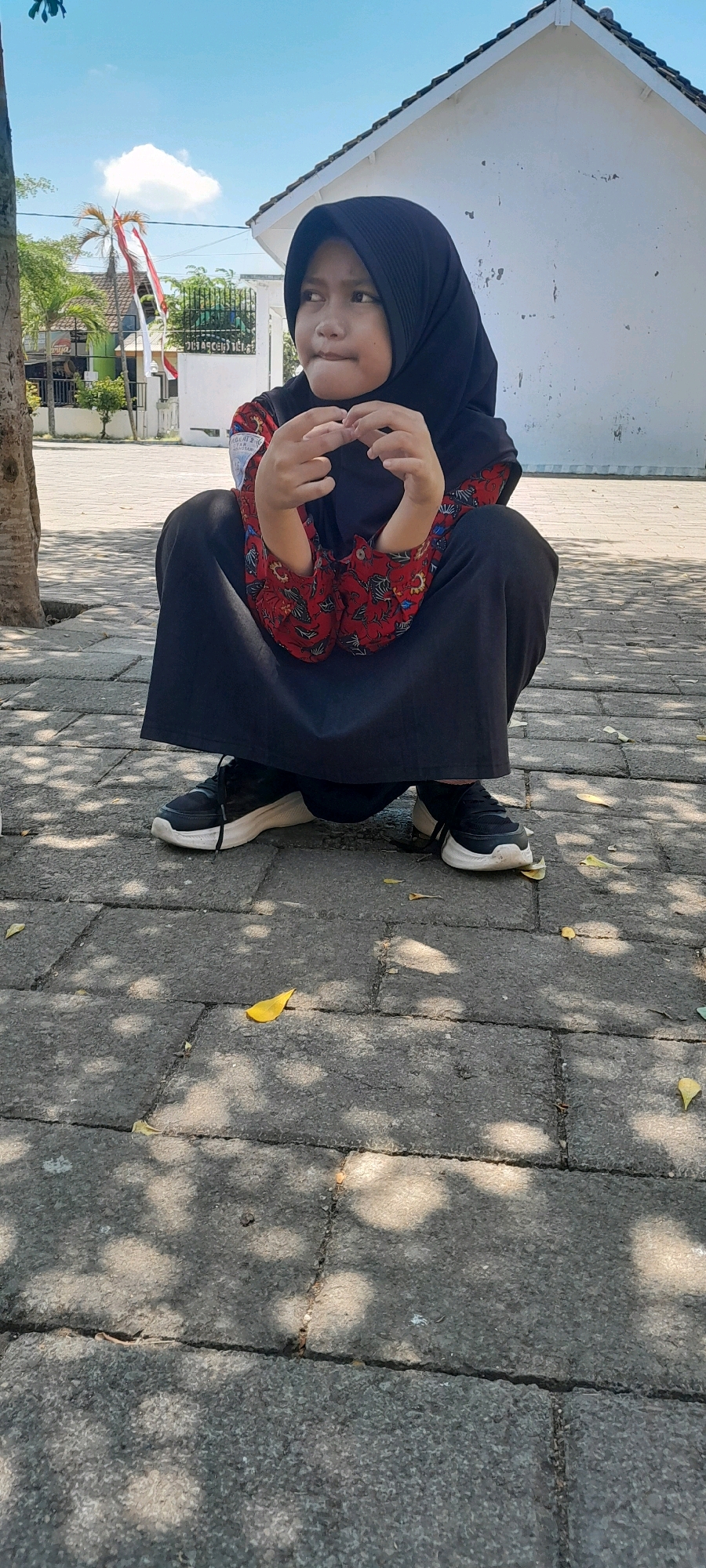 A young girl wearing a black hijab and a red and black patterned shirt is crouching on a gray paved walkway. She is looking to her right with her mouth slightly open and her hands are positioned in front of her chest, with her fingers interlocked. The ground is paved with rectangular gray stones, and dappled sunlight casts shadows from unseen foliage. In the background, a white building with a sloped roof is visible, along with some trees and a red and white flag. The scene appears to be outdoors in a public area, possibly a schoolyard or park, given the building and flag. The overall impression is a candid shot of a child in a moment of thought or play.
