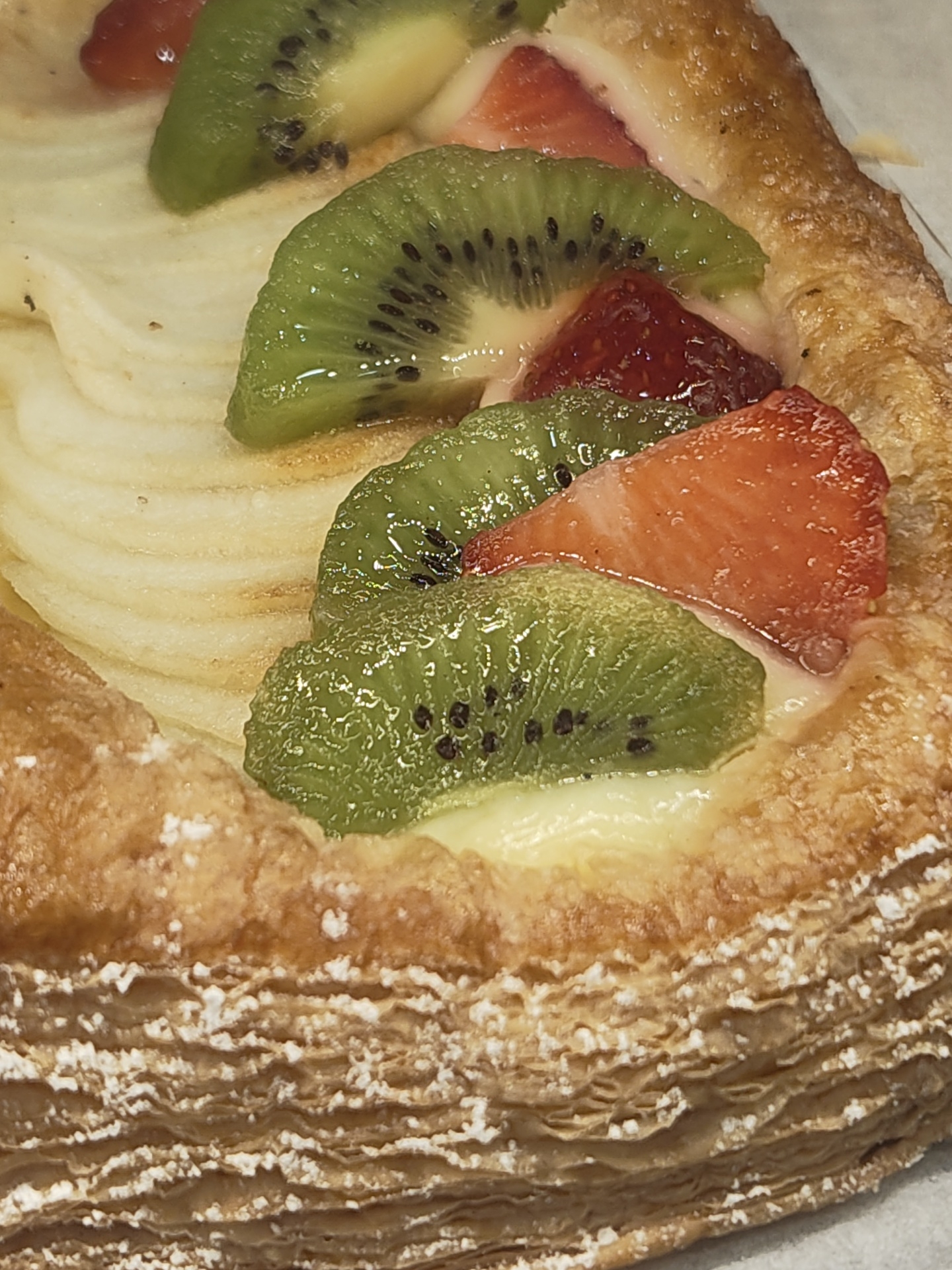 The image shows a close-up of a fruit pastry. It's topped with sliced kiwi, strawberries, and what appears to be thinly sliced apple or pear. The pastry is golden brown and flaky, with a dusting of powdered sugar along the edges. There is no indication that this image was taken in Dénia, Spain. There are no people, interactions, or any specific environmental markers visible to support that claim.