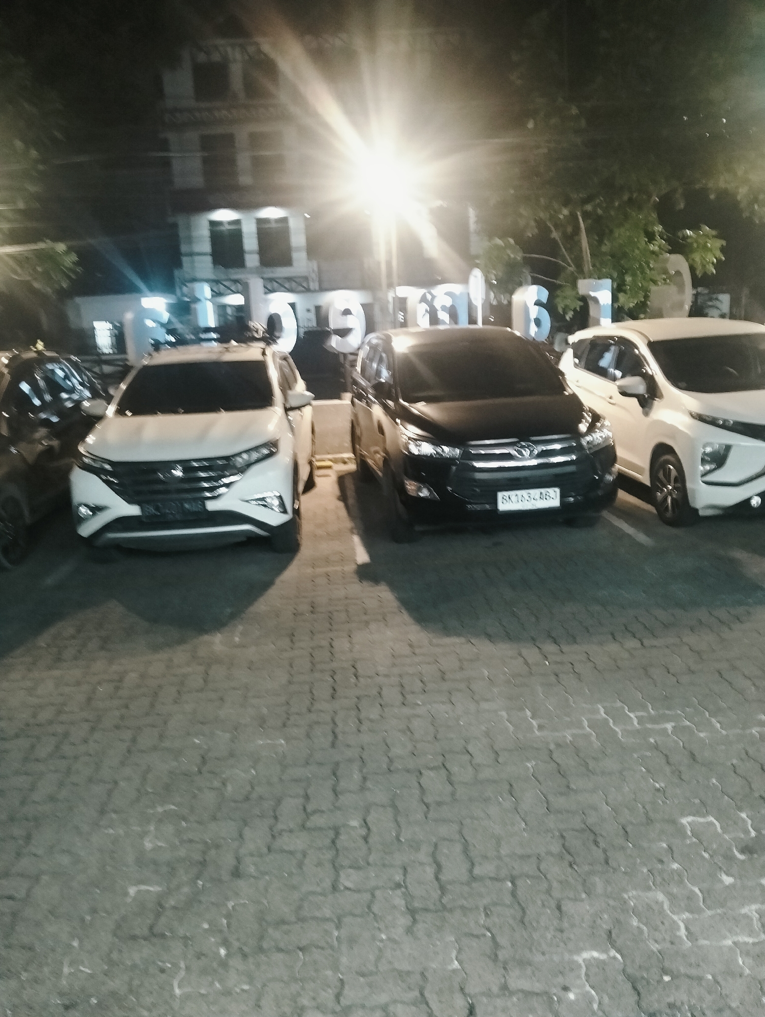In an outdoor, paved parking lot at night in the city of Medan, Indonesia, several parked vehicles are visible. A white SUV is in the foreground on the left, followed by a black SUV in the middle. To the right of the black SUV is a white car, and further to the right, another white car is partially visible. In the background, a building with multiple floors is illuminated, and large, illuminated numbers "16" are displayed. Trees are present behind the building. The scene is lit by bright overhead lights that cause lens flare.