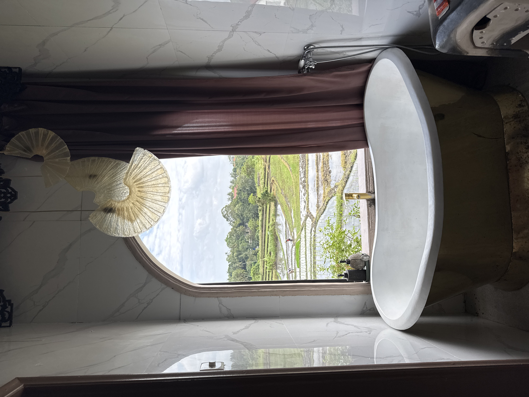 A freestanding bathtub with a gold exterior and white interior is positioned in front of an arched window overlooking lush green rice paddies with two figures working in them. The bathroom features marble-patterned walls and a dark brown curtain. Above the bathtub, a decorative light fixture with three fan-shaped shades hangs. On the windowsill, two soap dispensers and a small plant are visible. To the right of the bathtub, a metal object with a silver hose is attached to the wall. The scene is set in Mengwi, Indonesia.