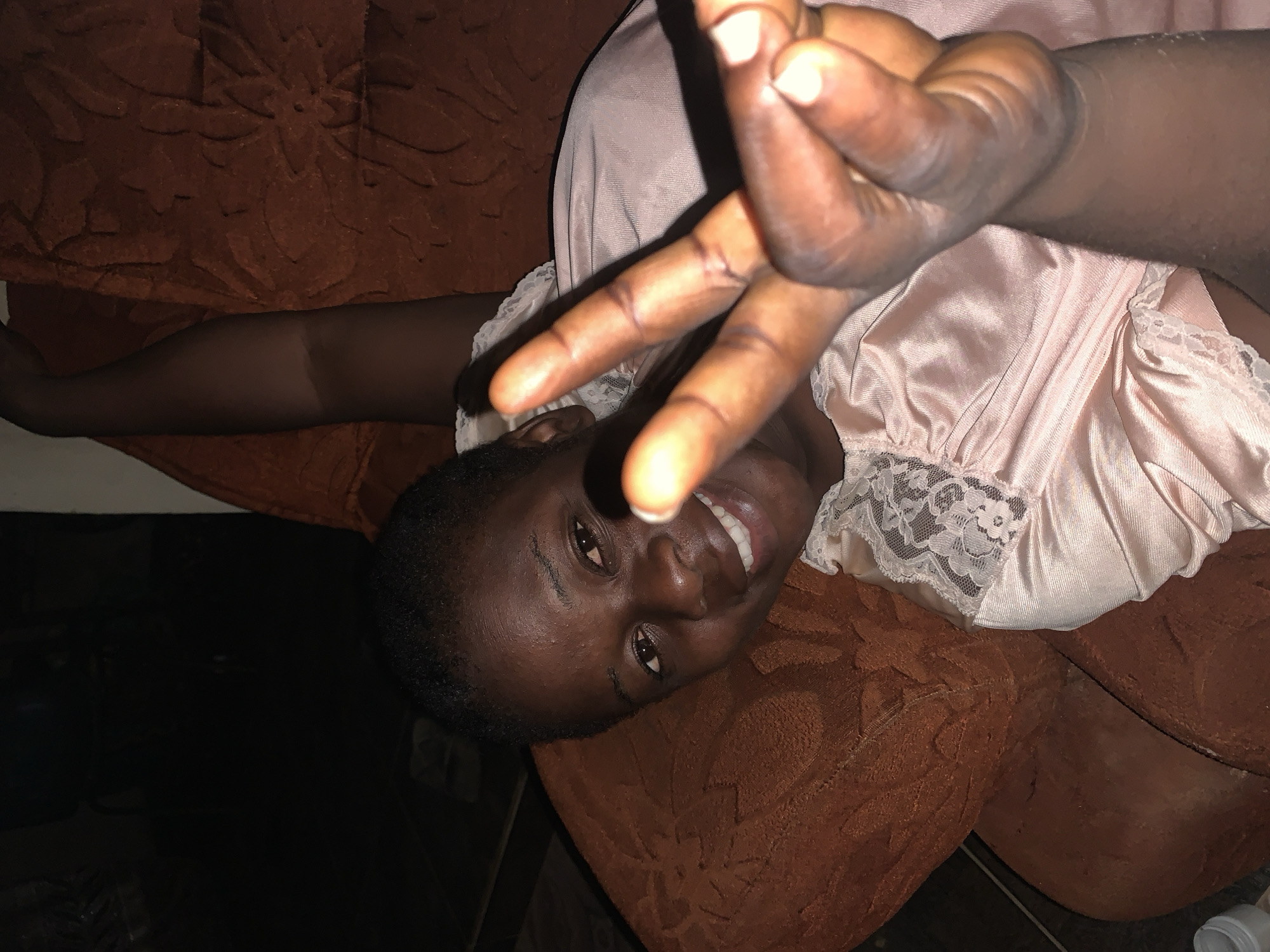 A woman is lying on a brown sofa, her head resting on the back cushion. She is smiling and making a peace sign with her right hand. She is wearing a light pink top with lace detailing. The location appears to be indoors. The city of Agban, Nigeria, cannot be determined from this image.