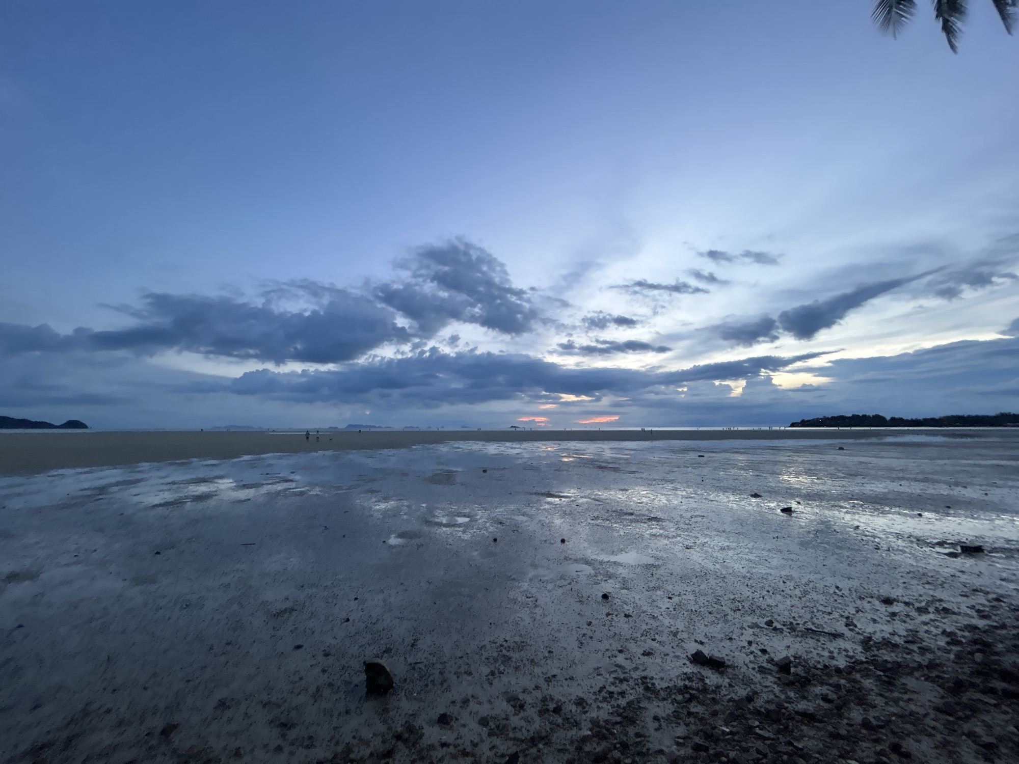 The image depicts a low-tide beach scene at dusk in Ko Pha Ngan, Thailand. The sky is a gradient of deep blue to lighter blue, with scattered, dark, and wispy clouds. In the distance, a thin strip of land with a few dark silhouettes of trees is visible. The foreground consists of wet sand, reflecting the sky and clouds. Small, dark rocks and debris are scattered across the sand. Several indistinct figures of people are present in the middle ground, walking on the exposed seabed. The overall mood is tranquil, with the dim light of twilight casting a serene atmosphere over the landscape.