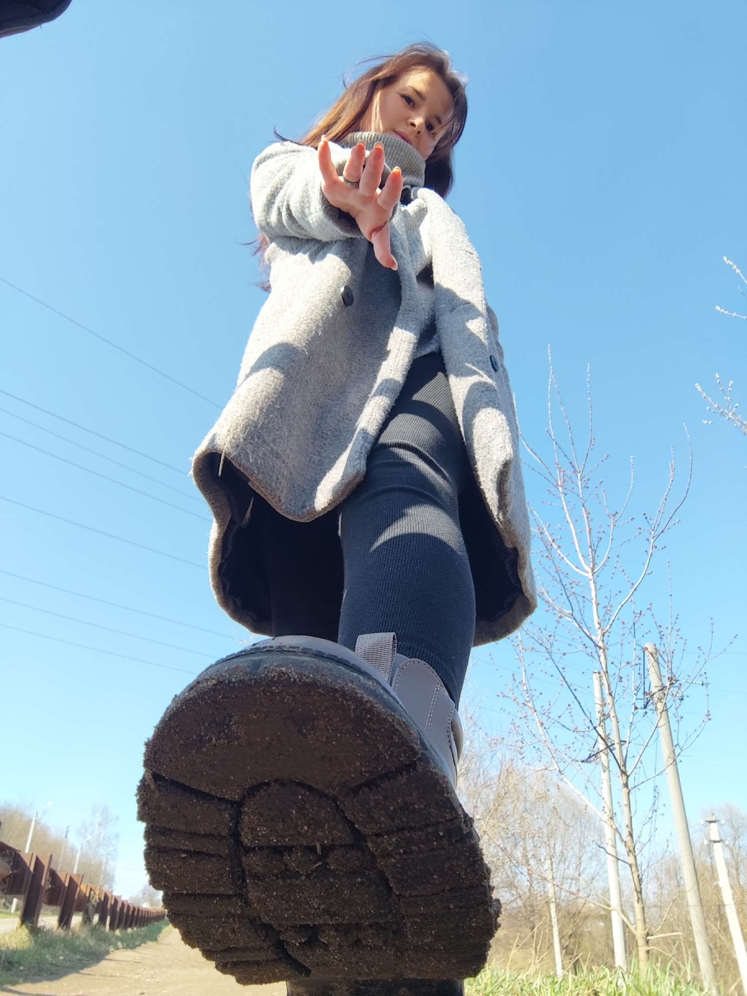 A woman stands in an outdoor area in Фоки, Russia. She wears a gray coat over a gray turtleneck and black leggings. Her right hand is outstretched toward the viewer. The bottom of her left shoe is the most prominent part of the photo, and it is covered in dirt. A metal guardrail and bare trees are in the background, under a clear, blue sky. Utility poles and wires are also visible in the background.