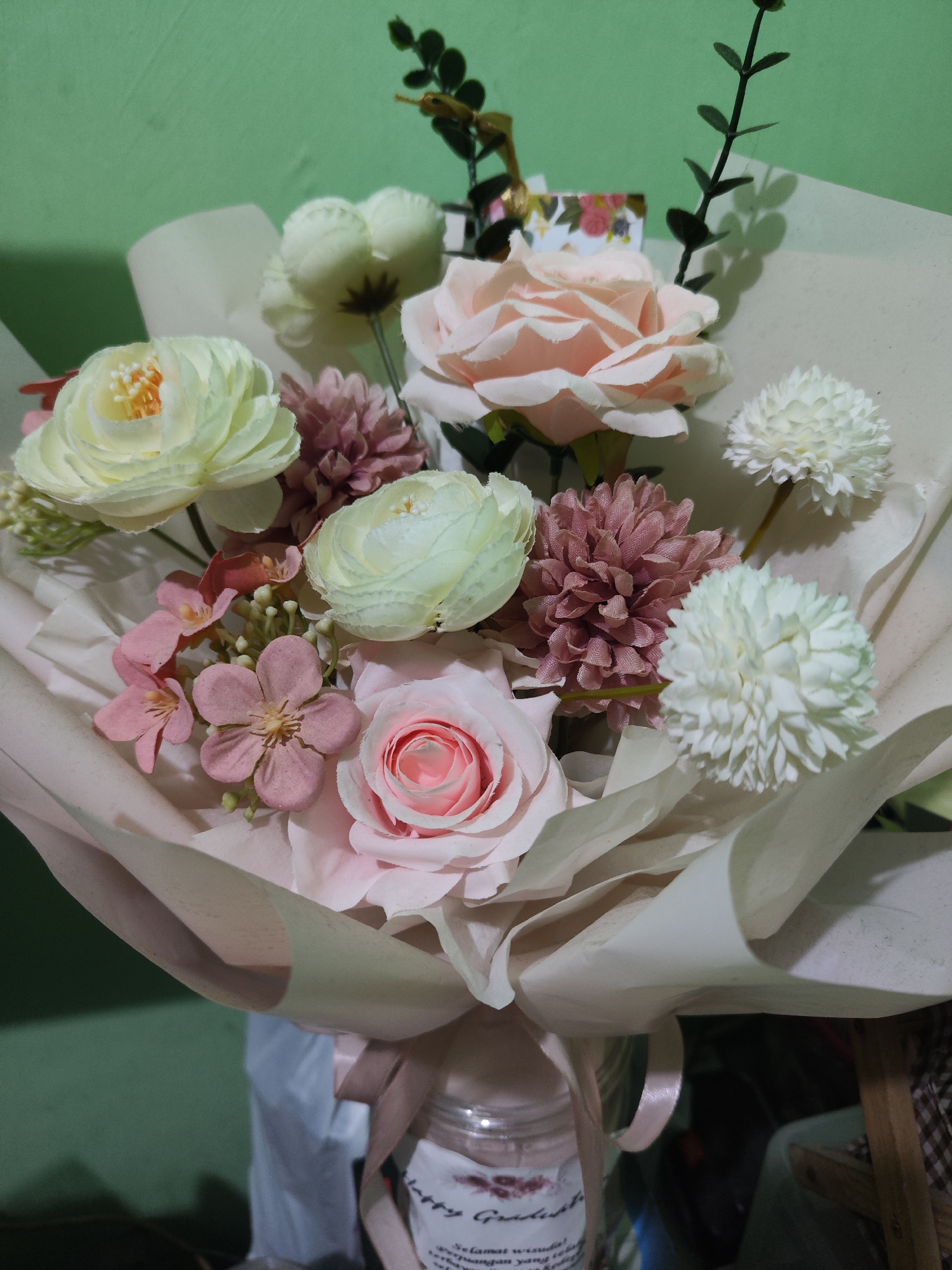 A bouquet of artificial flowers is displayed on a surface. The bouquet is composed of several types of flowers in shades of pink, white, and cream, including roses, chrysanthemums, and smaller, delicate pink flowers. These flowers are arranged with green foliage and are wrapped in light-colored paper, tied with a pink ribbon. The bouquet is held in a glass jar that has a label on it. The background is a plain green wall. The scene appears to be indoors, possibly in a room in Sroyo, Indonesia.