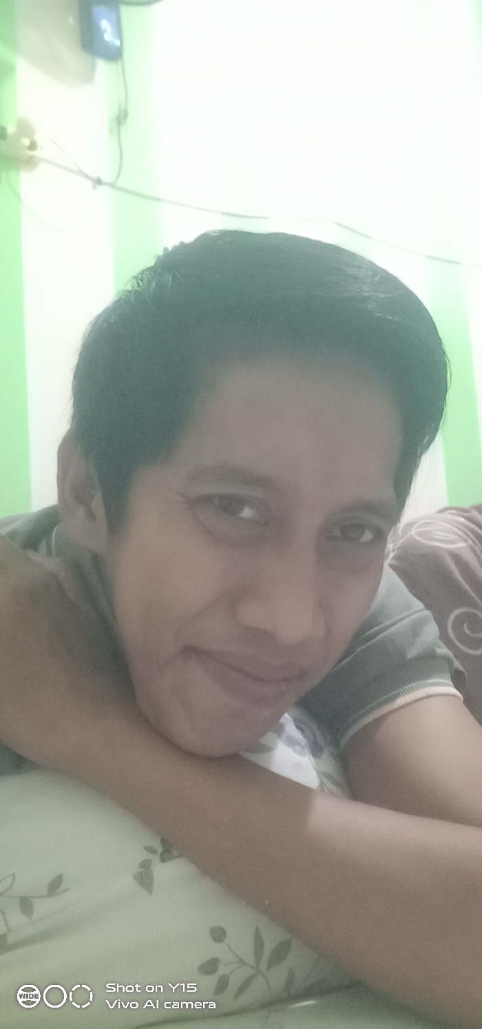 A medium close-up image depicts an adult male lying down, positioned in what appears to be a bedroom located in Ranugedang, Indonesia. The individual has short dark hair and a light complexion, looking directly at the viewer with a slight smile. He is wearing a grey short-sleeved collared shirt. His right arm is bent with the forearm supporting his head or chin, while his left arm is also bent, resting on light-colored bedding adorned with a subtle pattern of dark leaves and branches. To the subject's right, a brown or maroon blanket or pillow with a lighter circular motif is visible. The background features a wall painted with vertical alternating stripes of light green and white. In the upper left corner of the background, a white electrical outlet and a blue digital display unit, possibly an air conditioner control or clock, are mounted on the wall, with associated wiring visible. The scene is brightly illuminated.