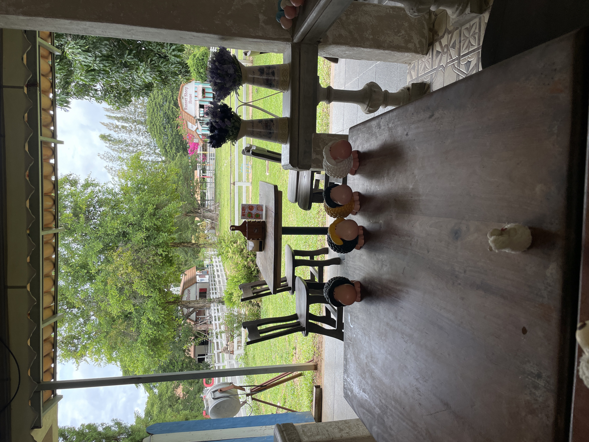 In Na Chom Thian, Thailand, this image captures a view from a covered patio looking out onto a farm-like setting. A wooden table and chairs are in the foreground, with decorative sheep figurines on the table. In the background, a white fence encloses a grassy area with trees and a small building labeled "Runner Sheep House." Two vases of lavender are displayed on a shelf. The scene suggests a relaxed outdoor dining or resting area.