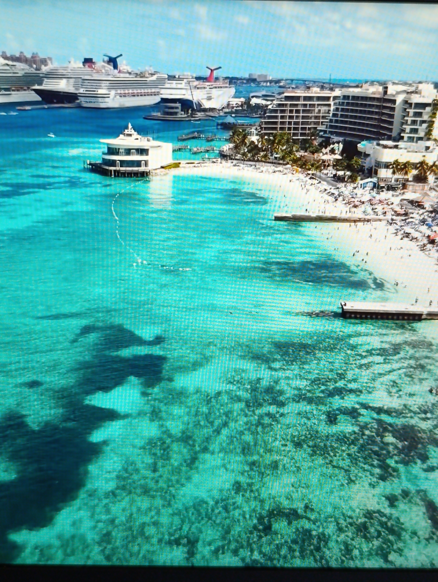 An aerial, high-angle view of a tropical coastline features crystal-clear turquoise water with visible coral reefs and sandy seabed. Several large cruise ships are docked in the distance. The foreground shows the shallow, clear water. A white, circular building with an overhang sits on a short pier extending into the water. Beyond this structure is a white sandy beach crowded with numerous sunbathers. Several palm trees are scattered along the beach, and modern, multi-story buildings with many windows line the shore behind the beach. Two piers extend from the beach into the water. The sky is bright and appears to be partly cloudy. The image is taken from a high vantage point, possibly an airplane or drone, looking down at the scene. The exact location in the city of Dubăsari town council, Moldova, is not depicted in this image.