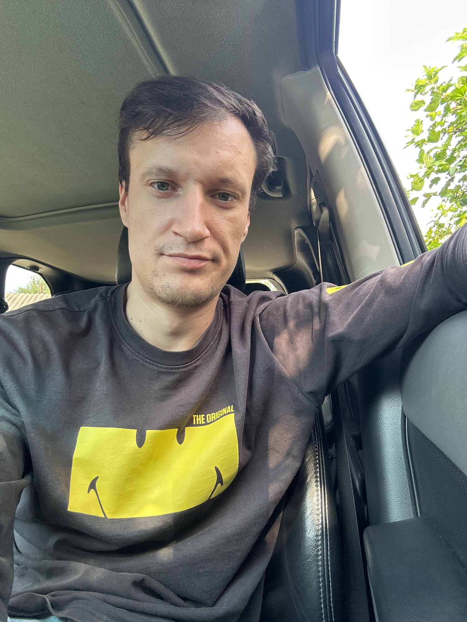 The image is a selfie of a man sitting in the driver's seat of a car. He is wearing a dark gray sweatshirt with a large yellow smiley face graphic and the words "THE ORIGINAL" written above it. The man has short brown hair and is looking towards the camera with a neutral expression. The interior of the car is visible, including the dashboard, sunroof, and a portion of the passenger seat. Sunlight is streaming into the car, casting shadows on the man and the interior. Outside the car, greenery and trees are visible through the windows, suggesting an outdoor, possibly rural or suburban, setting. The location is specified as Shatava, Ukraine.