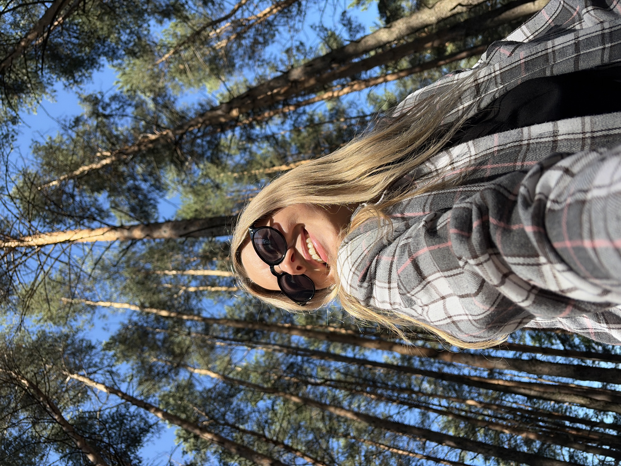 A low-angle, close-up, outdoor, selfie-style photograph shows a young woman with blonde hair and sunglasses smiling at the camera. She is wearing a grey, white, and pink plaid shirt over a black top. The background is a forest with tall, slender trees reaching towards a clear blue sky. The image appears to have been taken in Chaikovsky, Russia, based on the context provided.