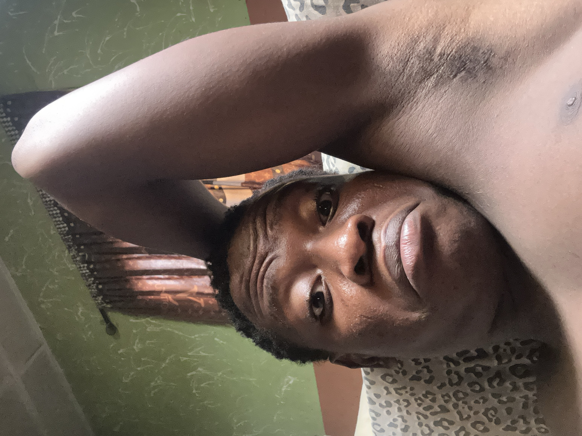 The image features a close-up, high-angle view of an adult male with dark skin, lying down and looking directly at the camera. His head is positioned towards the right side of the frame, and his body extends diagonally towards the upper left.
The man appears bare-chested, with his right arm bent and raised above his head, partially visible in the upper left quadrant. He has a neutral to slightly pensive expression, characterized by a slightly furrowed brow and slightly parted lips.
The background on the left side of the frame consists of a greenish wall with subtle white, swirling patterns. Behind his head, in the middle-left background, a window with light brown wooden blinds is visible. On the right side of the frame, a patterned fabric, resembling leopard print, can be seen, likely part of bedding or a garment. Notable details include the visible texture of his skin, some short dark hair in his armpit and on his arm, and the soft lighting that highlights his features. There is no visible text in the image.