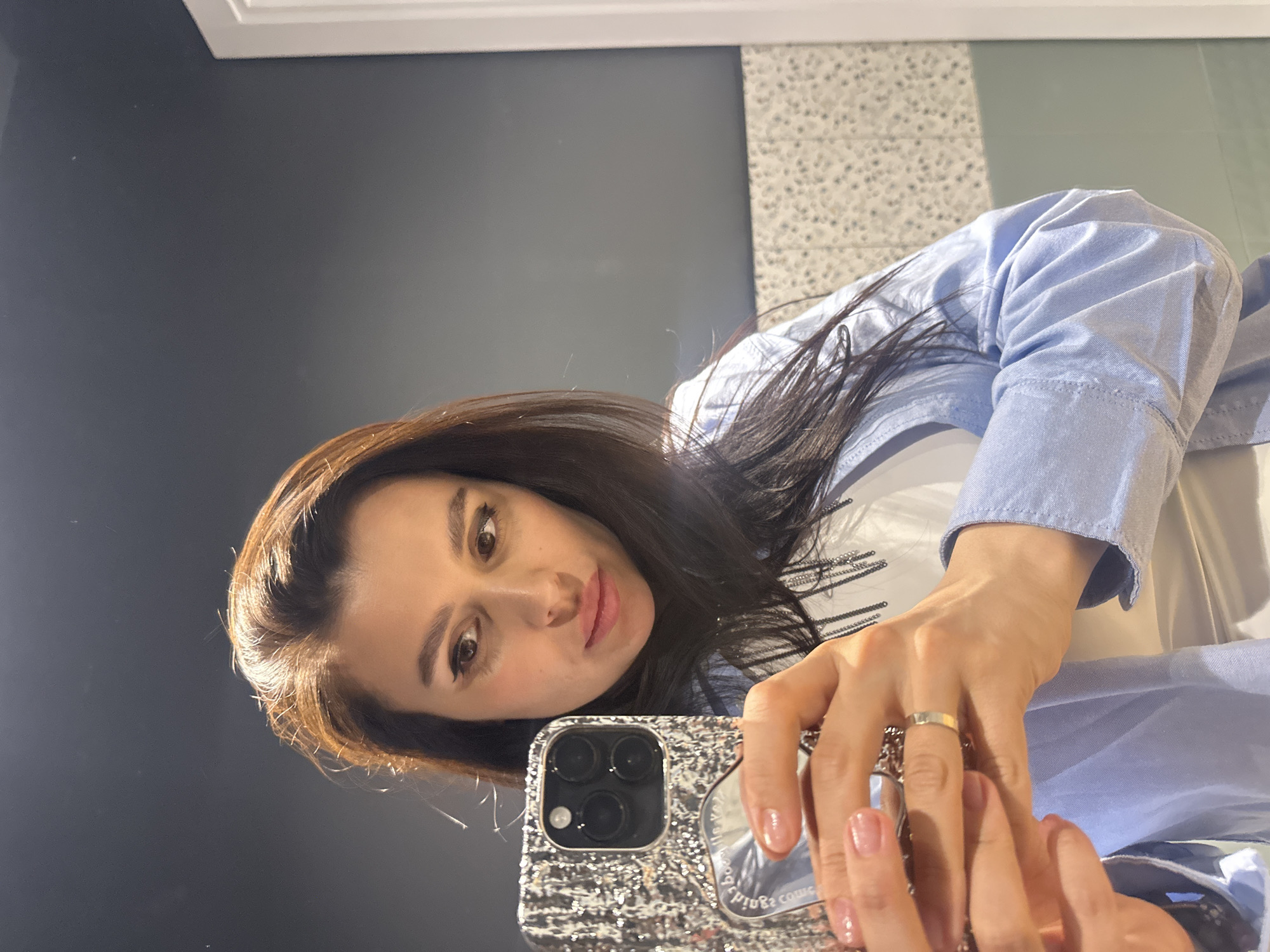 A woman is taking a selfie in what appears to be a bathroom. She is holding a smartphone with a textured silver case. The woman has long, dark hair and is wearing a light blue button-down shirt over a white shirt. A gold ring is visible on her finger. The background includes a gray wall on the left and a wall with a speckled pattern on the right. I have no information about the location in the city of Chaikovsky, Russia.