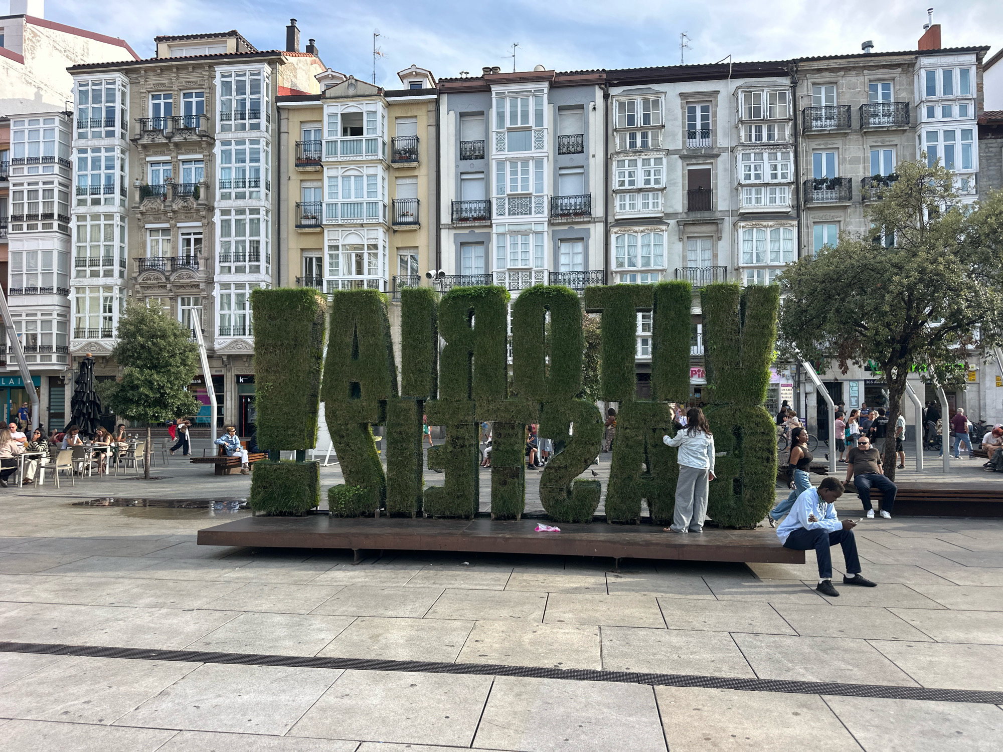 In Vitoria-Gasteiz, Spain, a large, green, sculpted hedge spelling out "LA GRANDEZA" stands prominently in a paved public square. The letters are three-dimensional and sit on a dark wooden platform. In the background, a row of multi-story buildings with intricate architectural details, including bay windows and balconies, line the square. The sky is partly cloudy. Several people are visible in the scene: a person wearing a light blue jacket and grey pants stands by the hedge, interacting with it; another person in a light blue sweatshirt is seated on the wooden platform, looking at a mobile phone; and further in the background, other individuals are seated at tables outside a cafe, walking, and sitting on benches. The overall atmosphere suggests a bright, daytime urban setting.